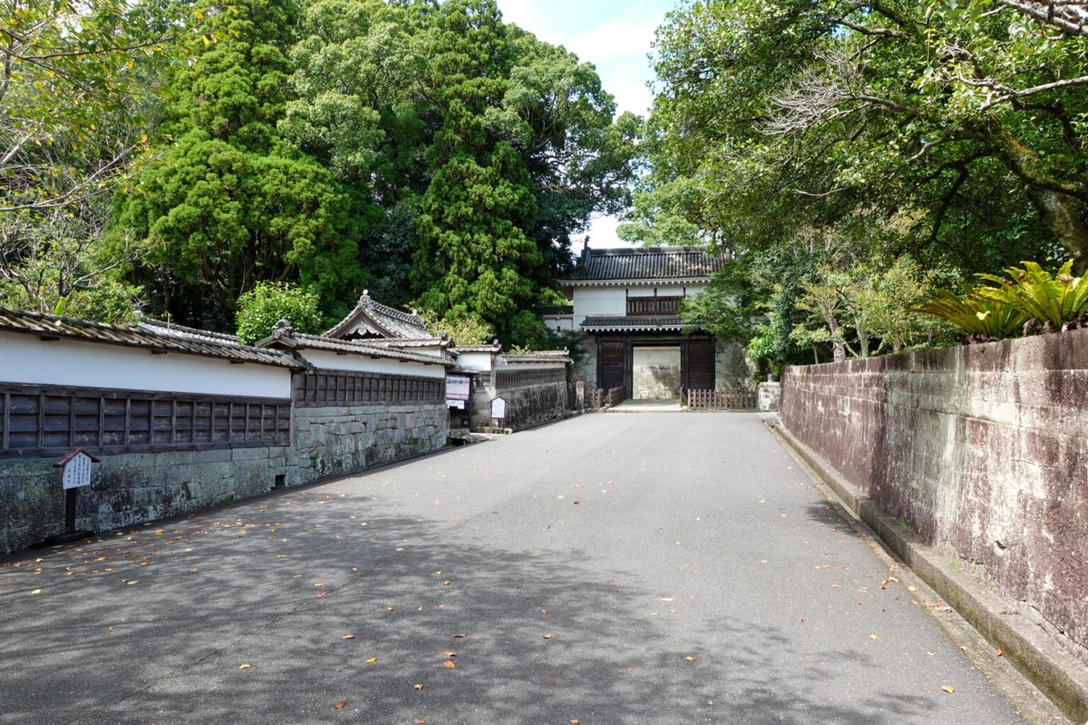 The broad approach road to Obi Castle, flanked by white samurai-residence walls on one side and a stone embankment on the other, with a white turret gate visible ahead among tall trees.