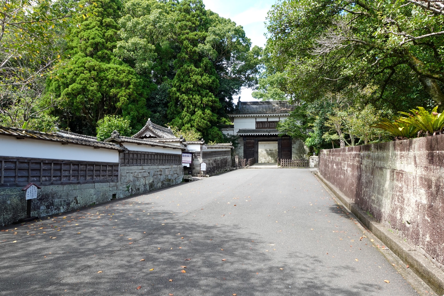The broad approach road to Obi Castle, flanked by white samurai-residence walls on one side and a stone embankment on the other, with a white turret gate visible ahead among tall trees.