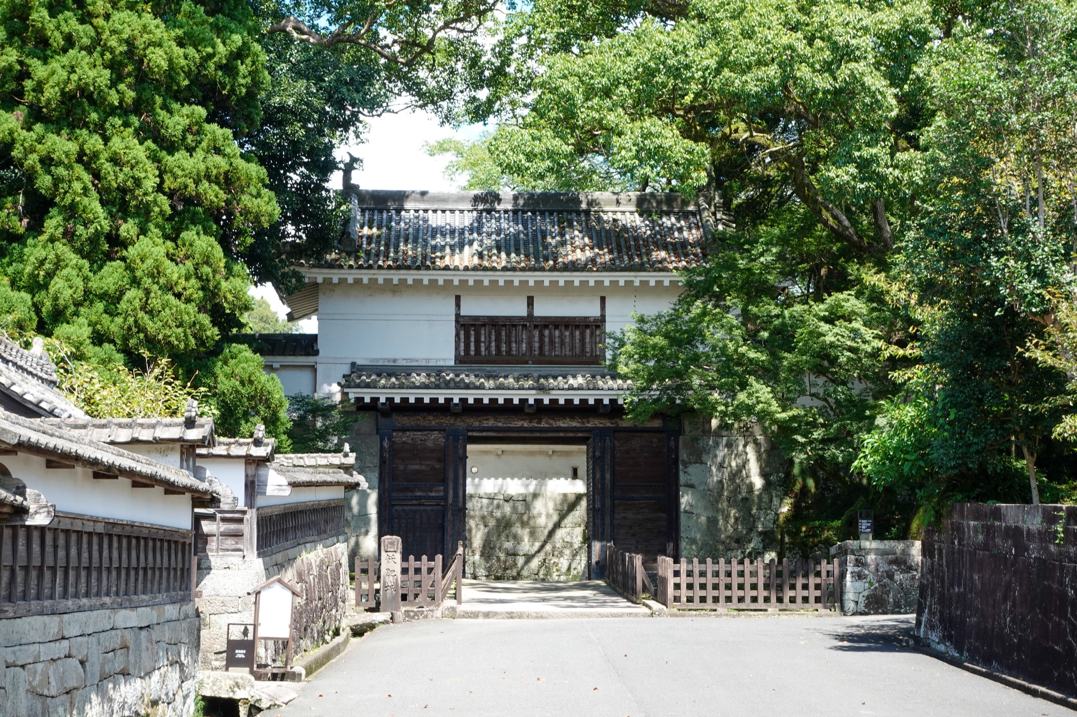 The reconstructed wooden Otemon main gate of Obi Castle in Nichinan, Miyazaki, a two-storey white-plastered turret gate above heavy black timber doors, framed by trees and the castle's old stone walls.