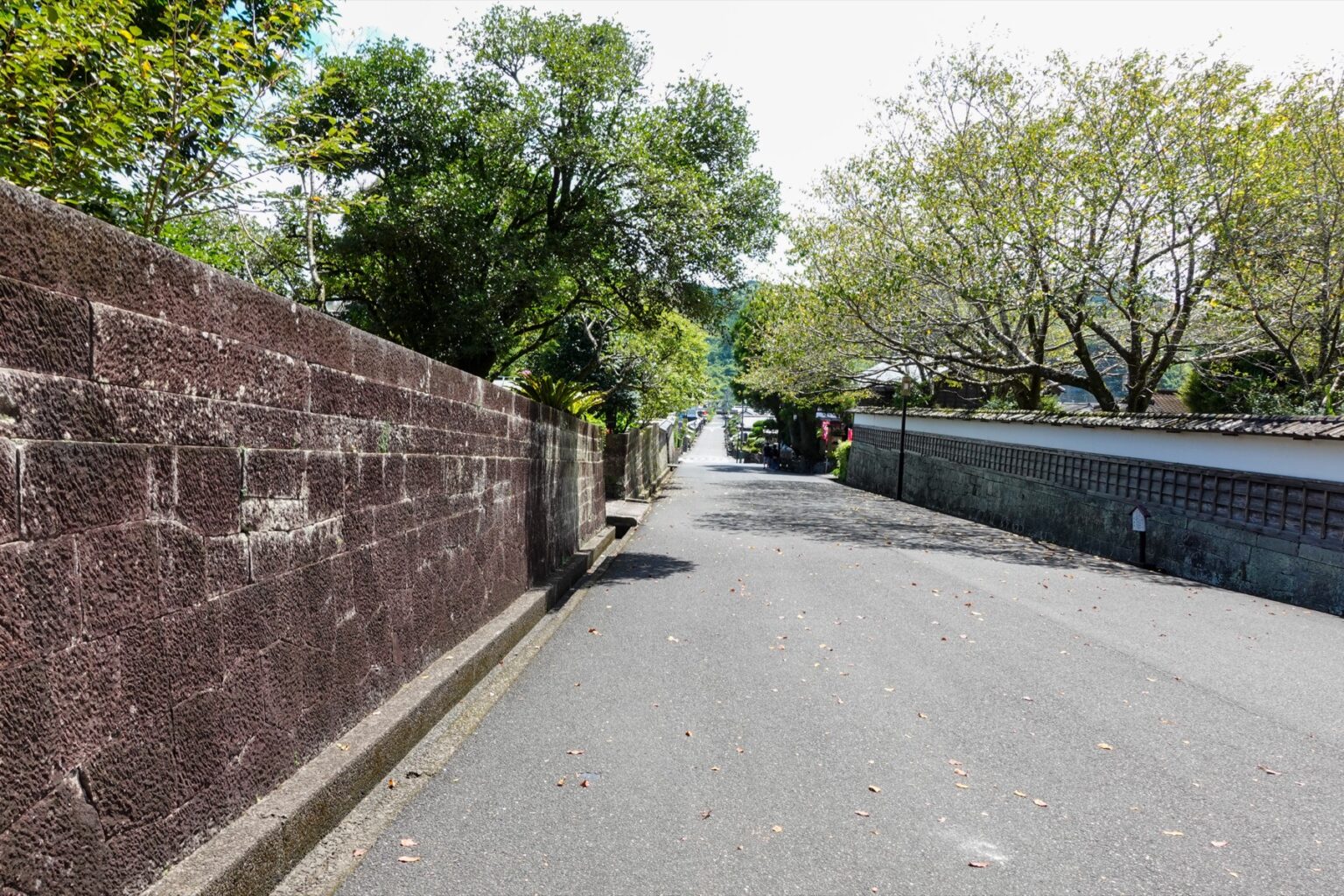 A quiet street in Obi's former samurai district, lined on the left by a long wall of reddish Obi stone and on the right by white plastered walls and trees, sloping gently uphill under a bright sky.