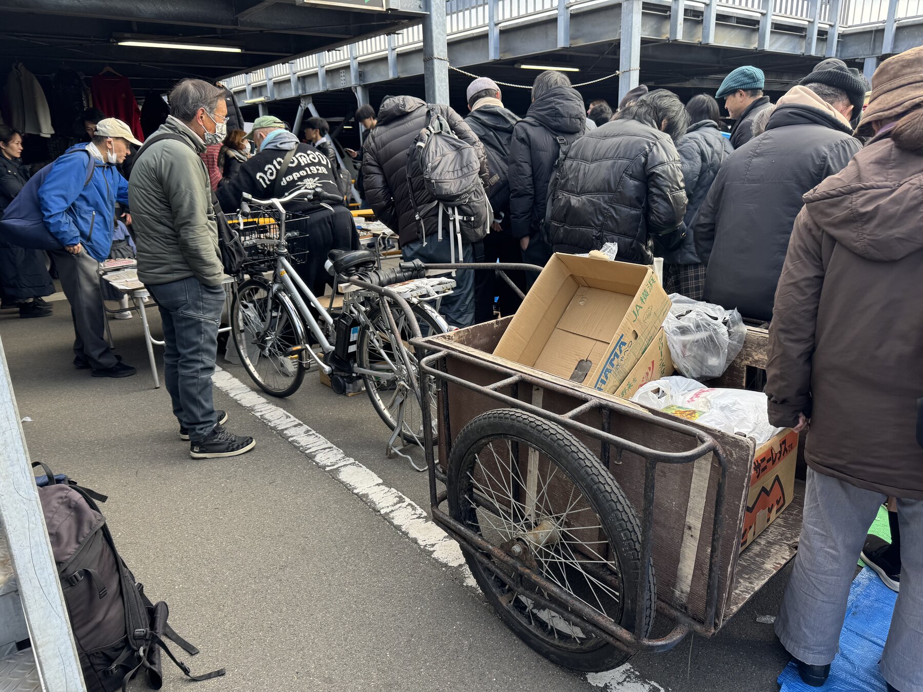 Shoppers and a vendor with a bicycle and a hand-cart loaded with goods along an aisle of the Ōi Racecourse flea market in Tokyo.