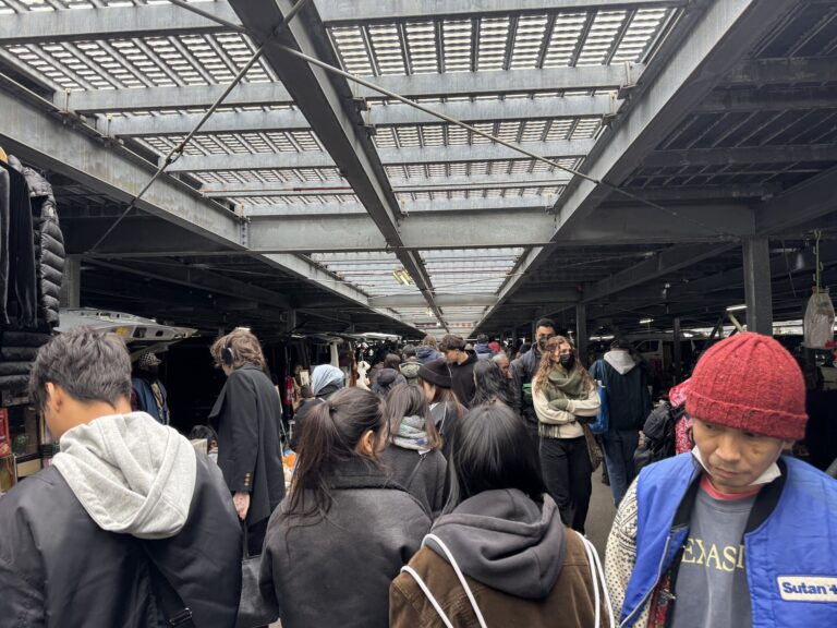 Crowds of shoppers browsing rows of stalls under the covered parking deck at the Oi Racecourse flea market in Tokyo, racks of clothing and tables of goods lining both sides.