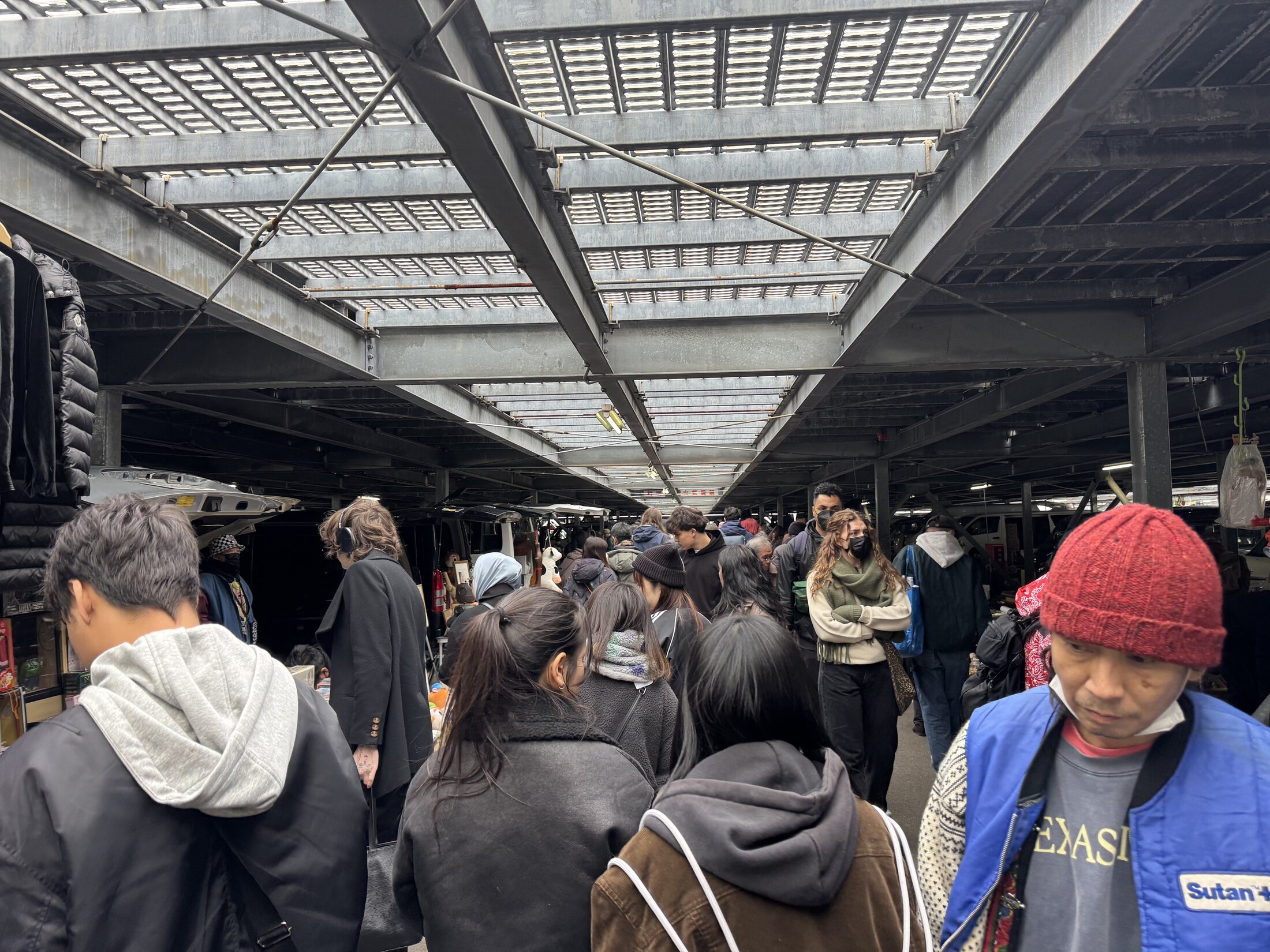 Crowds of shoppers browsing rows of stalls under the covered parking deck at the Oi Racecourse flea market in Tokyo, racks of clothing and tables of goods lining both sides.