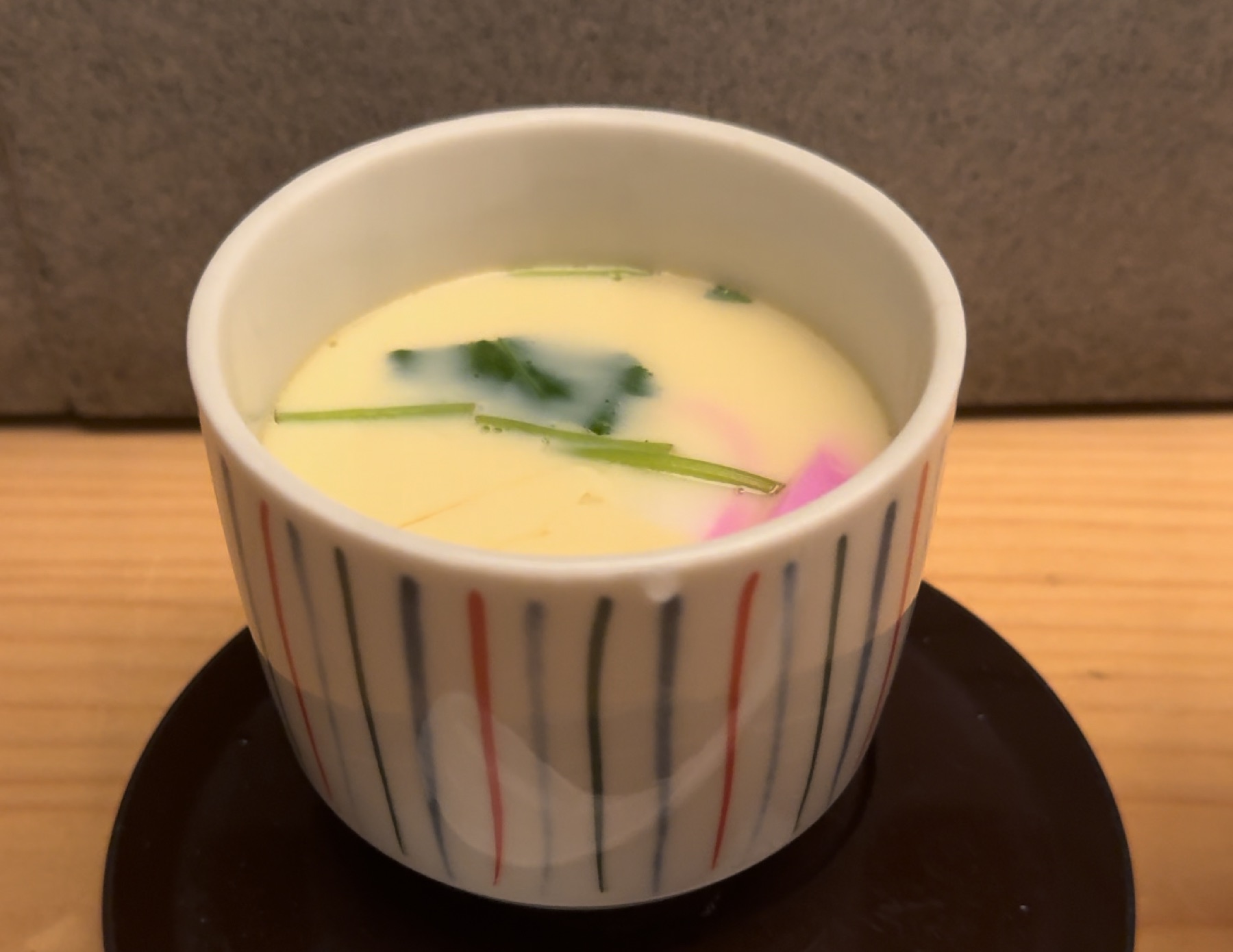 A chawanmushi egg custard in a hand-painted striped cup on a dark lacquer saucer at Orizuru in Kinosaki Onsen, garnished with green mitsuba leaves and a slice of pink-and-white kamaboko fish cake just visible under the surface.