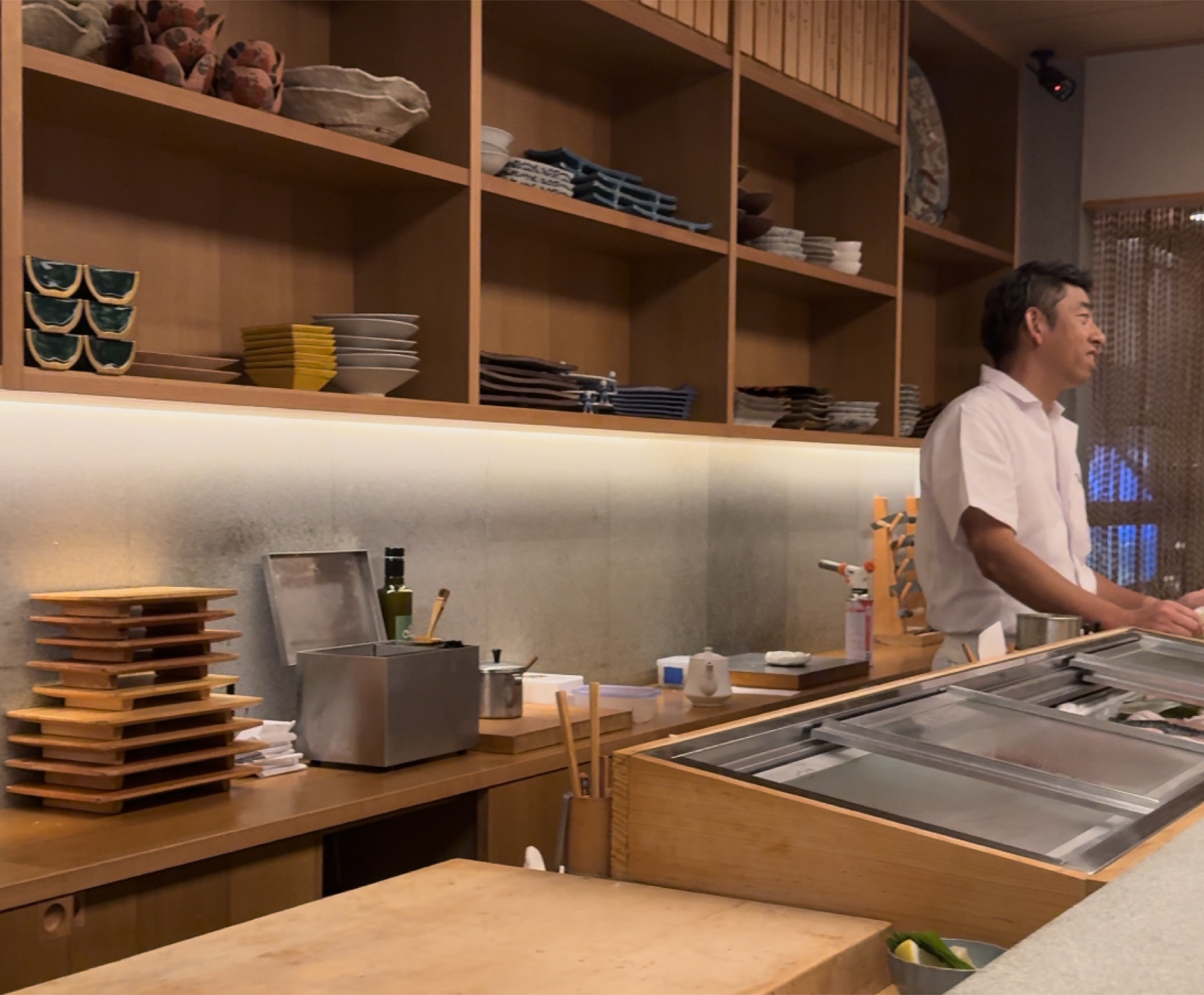 The wooden sushi counter inside Orizuru in Kinosaki Onsen, with the chef in a white shirt standing behind a refrigerated fish case and wooden shelves stacked with ceramic plates, bowls and cups in the background.