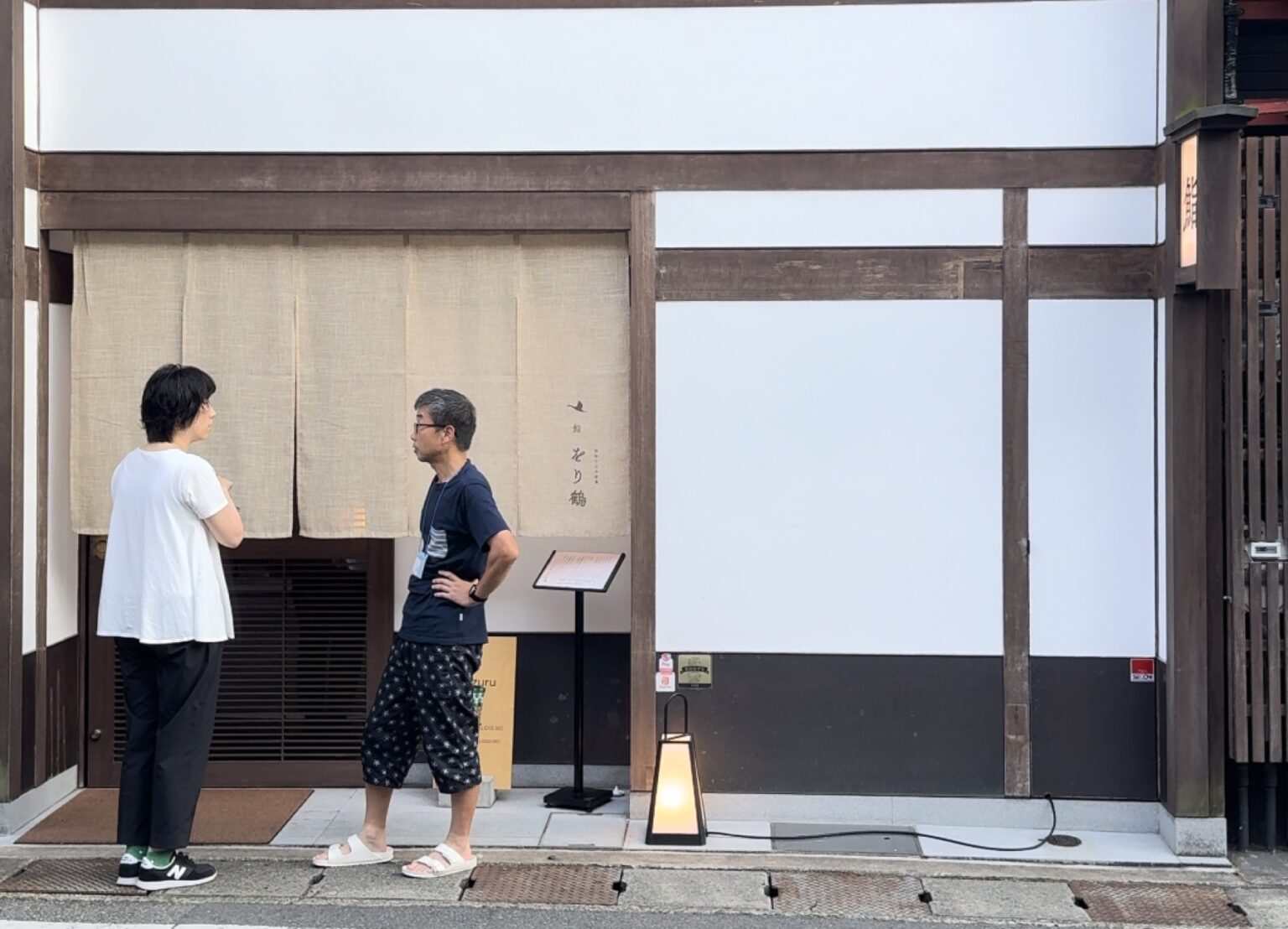 The street frontage of Orizuru sushi restaurant in Kinosaki Onsen, a machiya-style building with a beige fabric noren curtain printed with a crane mark, a standing menu board and a paper lantern, where two people stand talking outside the entrance.
