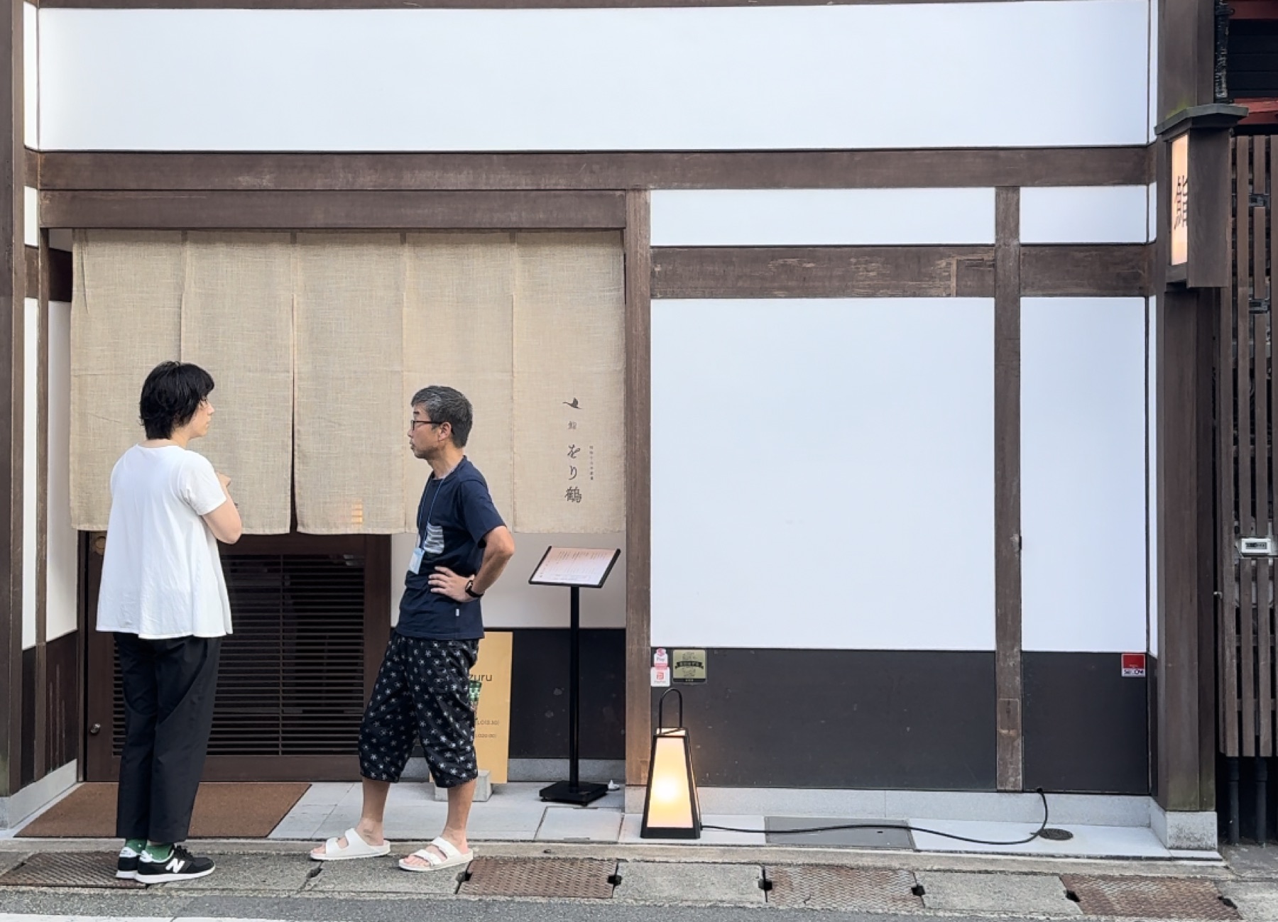 The street frontage of Orizuru sushi restaurant in Kinosaki Onsen, a machiya-style building with a beige fabric noren curtain printed with a crane mark, a standing menu board and a paper lantern, where two people stand talking outside the entrance.
