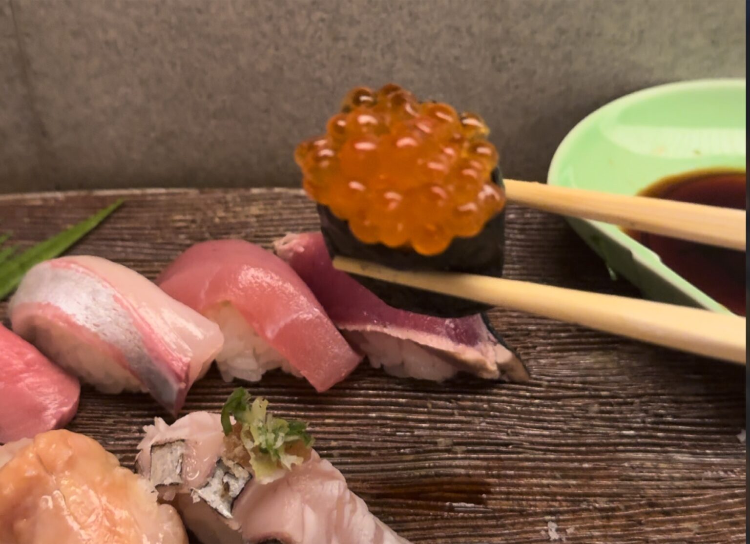 A close-up at the Orizuru counter in Kinosaki Onsen of a salmon-roe gunkan piece lifted with chopsticks above a row of nigiri, with a small dish of soy sauce on the right.