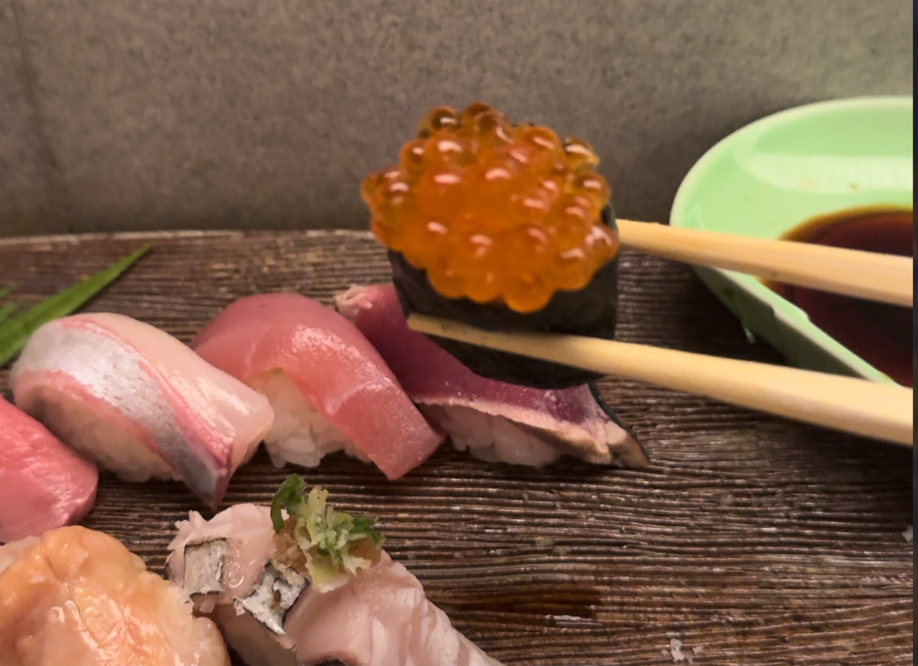 A close-up at the Orizuru counter in Kinosaki Onsen of a salmon-roe gunkan piece lifted with chopsticks above a row of nigiri, with a small dish of soy sauce on the right.