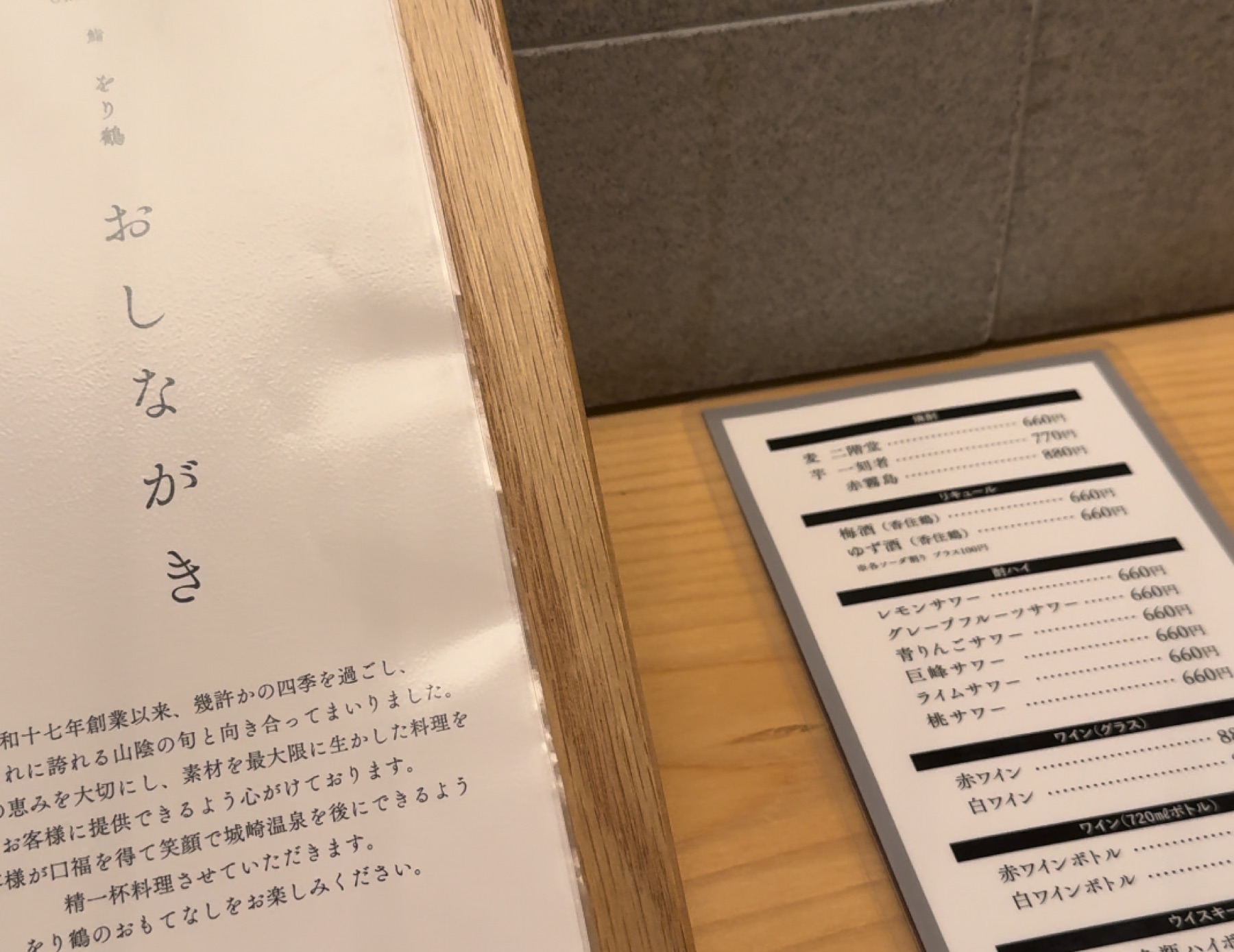 The oshinagaki menu cover at Orizuru in Kinosaki Onsen, printed with the restaurant name and a paragraph noting it has served seasonal San-in ingredients in Kinosaki Onsen since Showa 17, with a laminated drinks price list resting on the wooden counter beside it.