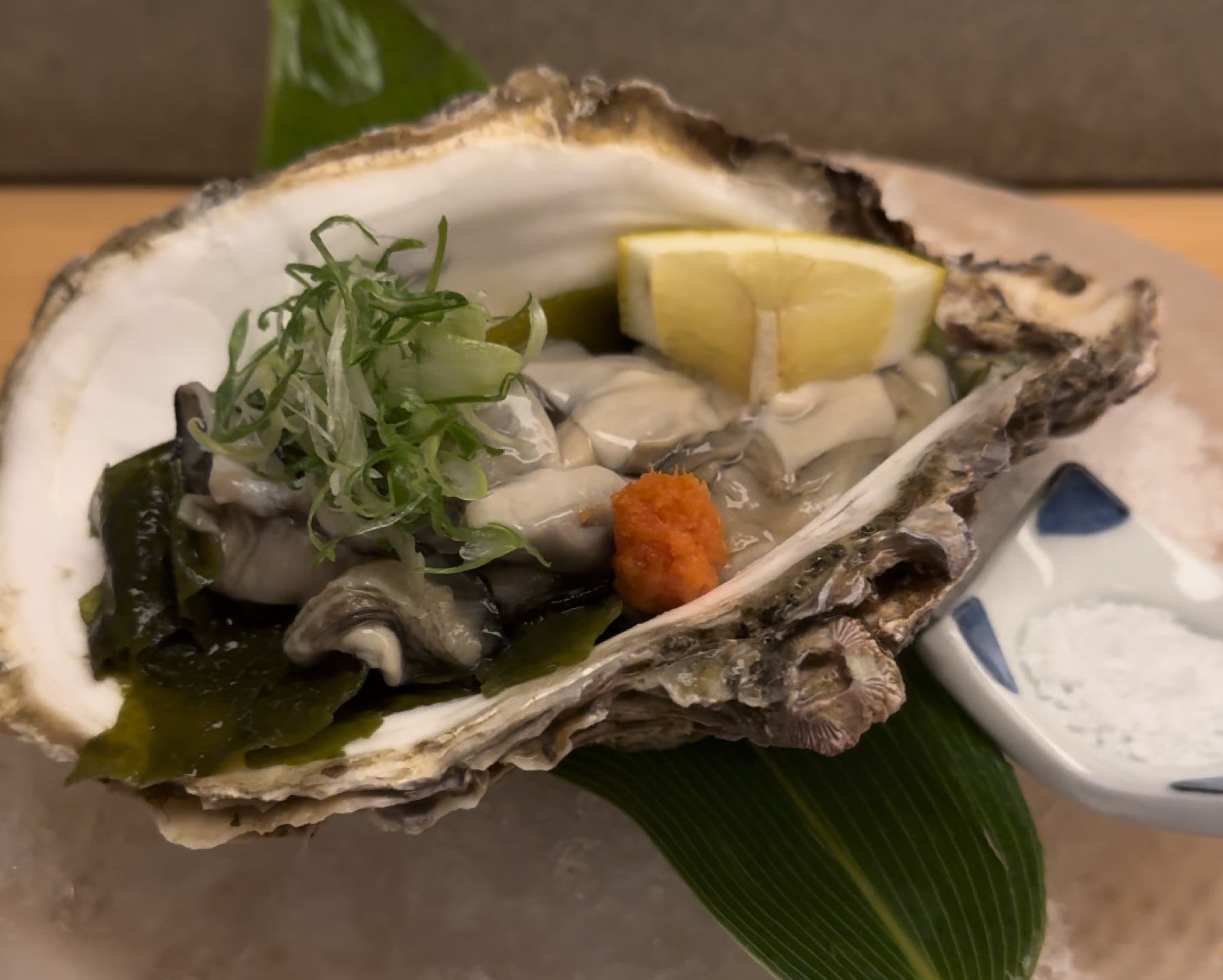 A single oyster served on the half shell at Orizuru in Kinosaki Onsen, topped with shredded green onion and wakame seaweed, with a lemon wedge and a small mound of red grated-daikon chili to the side, and a small condiment dish set alongside on ice.