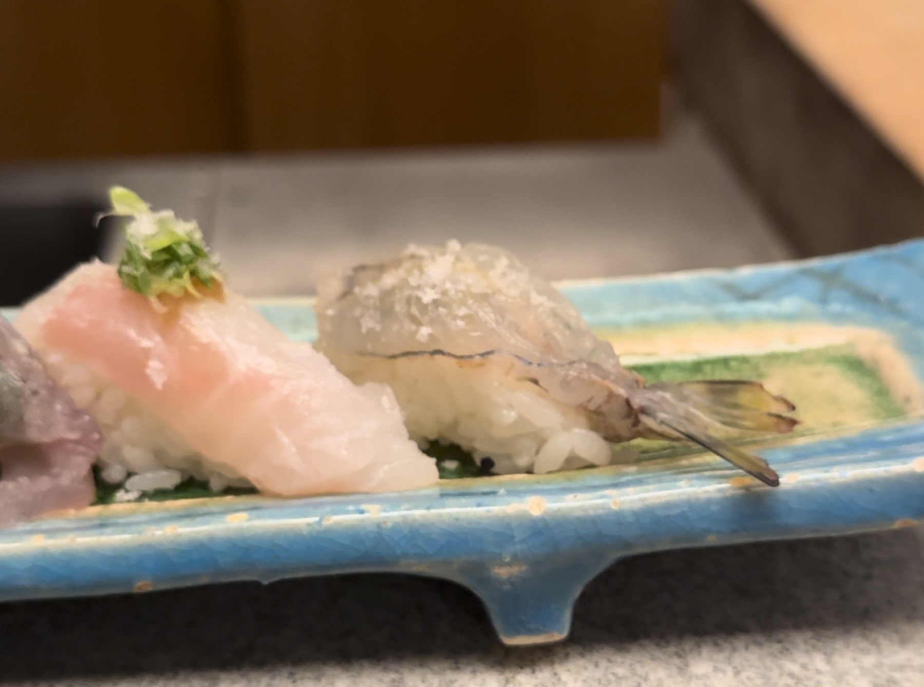 Two nigiri on a blue rectangular plate at Orizuru in Kinosaki Onsen: a white-fish piece topped with shredded green onion, and a translucent shrimp nigiri with the tail still attached, both resting on a green bamboo leaf.