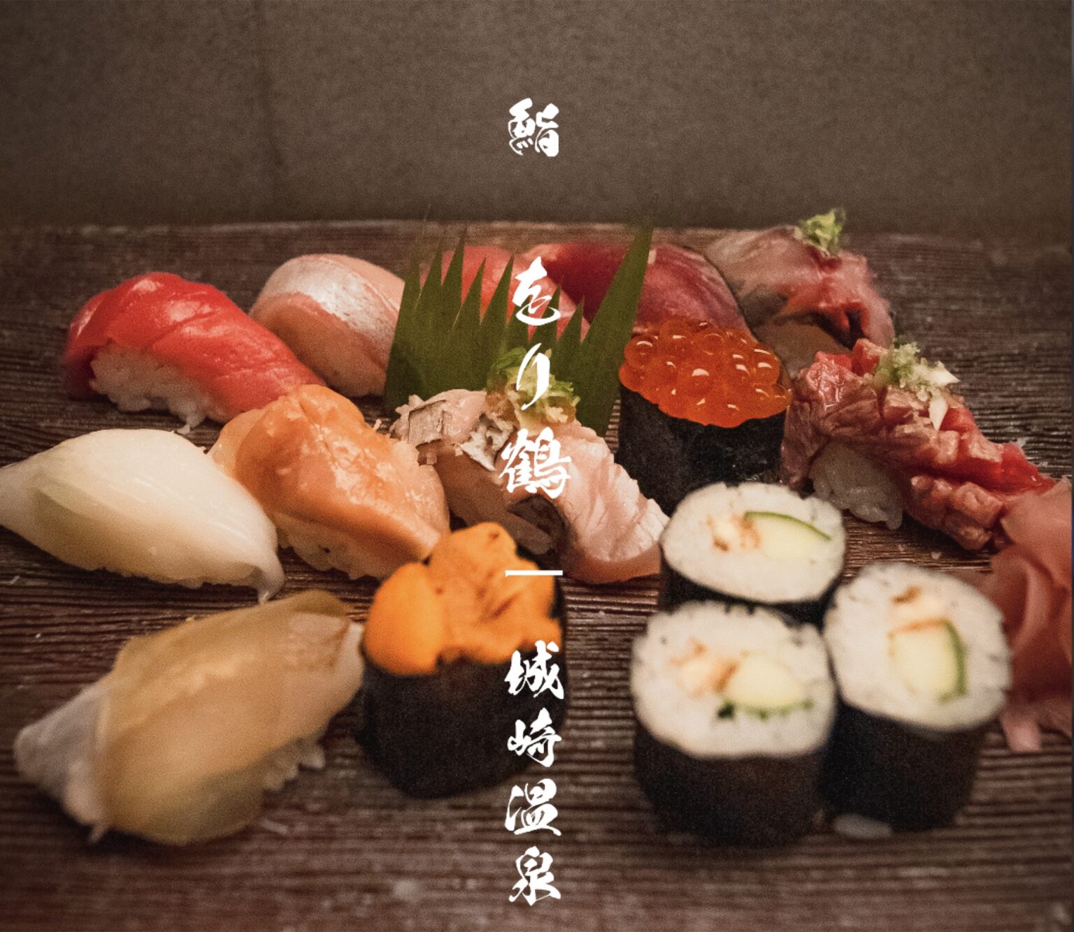 A wide assortment of nigiri, gunkan, and maki sushi arranged on a dark wooden board at Orizuru in Kinosaki Onsen, with vertical white Japanese lettering reading Sushi Orizuru Kinosaki Onsen.