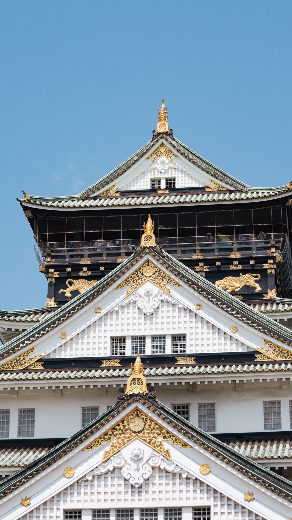 Detail of the upper floors of Osaka Castle's keep, with ornate white gables edged in gold leaf, golden tiger reliefs, and the open top-floor observation gallery where visitors look out over the city.