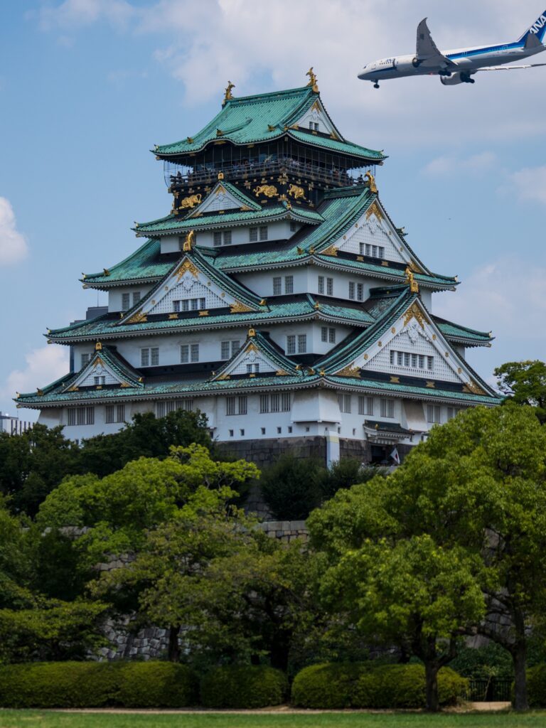 The reconstructed main keep of Osaka Castle rising above green trees, with white walls, green copper roofs and gold-leaf trim, as an ANA passenger jet passes low in the blue sky above the top floor.