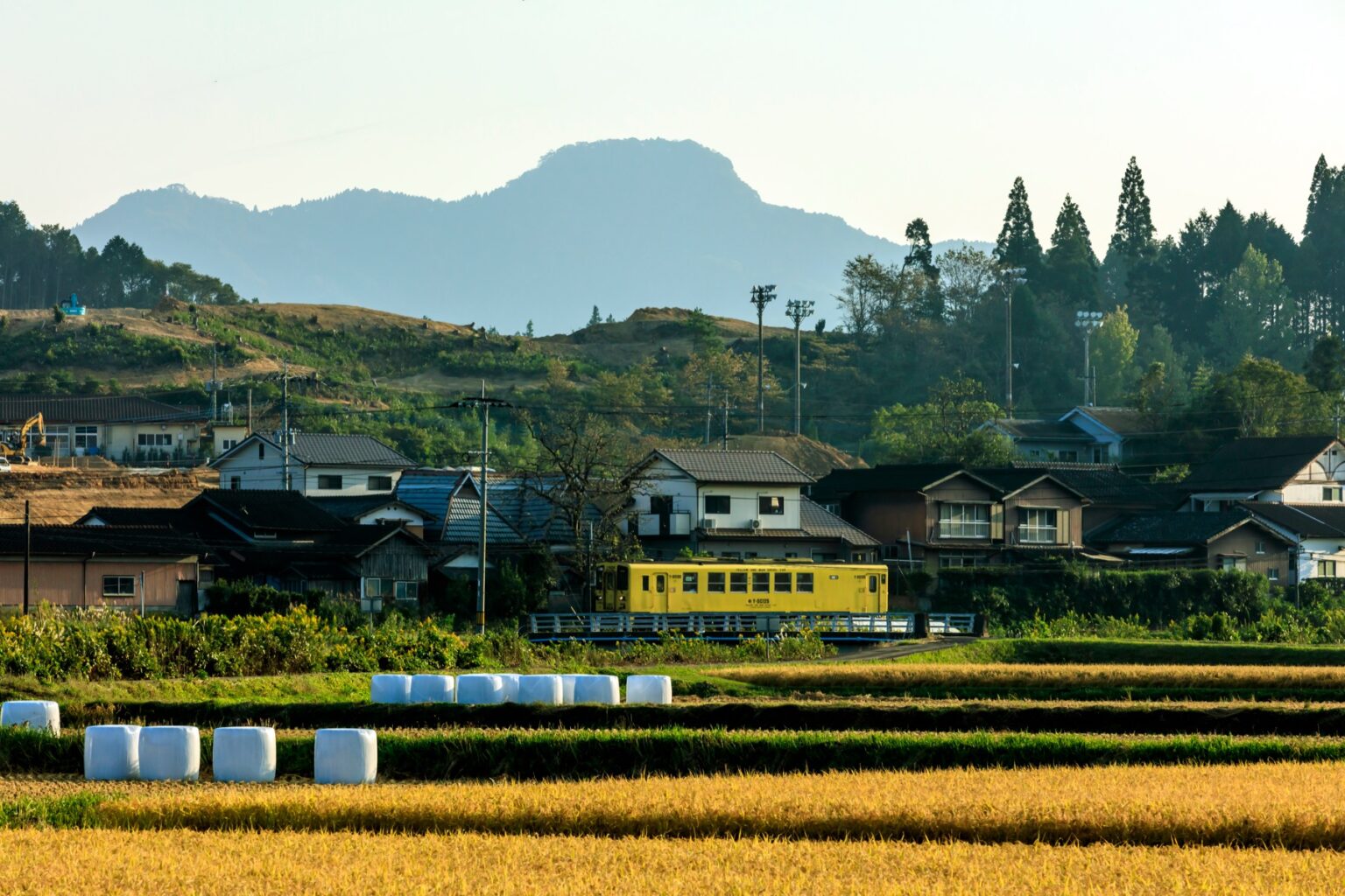 A yellow local train passing rice fields in rural Saga with mountains behind
