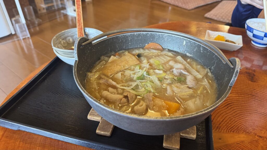 A black iron pot of pork hoto at Shoya by Lake Yamanaka: flat, wide wheat noodles simmered with pork, pumpkin and vegetables in a thick miso broth, on a wooden tray with rice and pickles