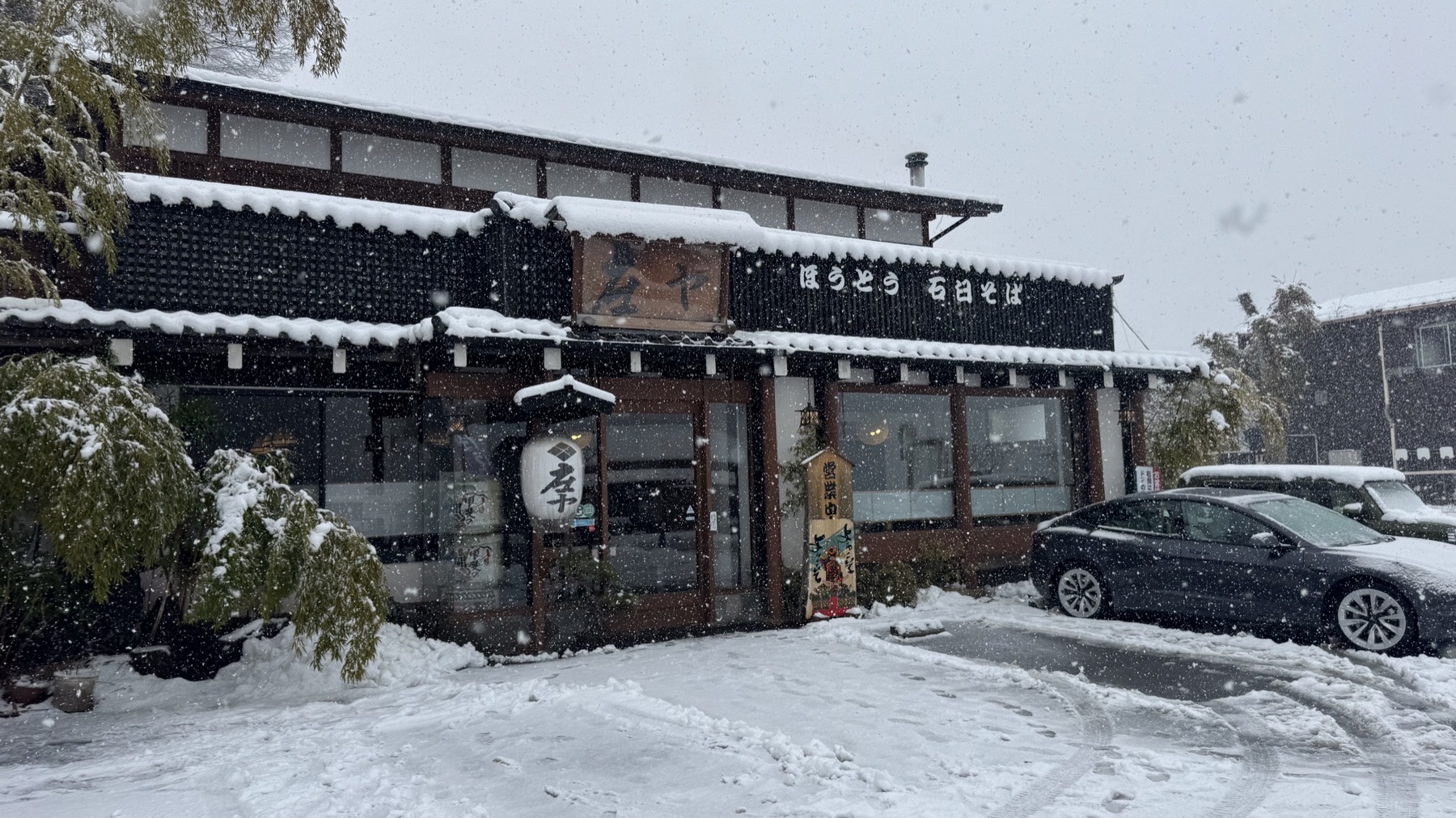 The wooden exterior of Shoya hoto restaurant by Lake Yamanaka in falling snow, with signs reading hoto and stone-milled soba and a car on the snow-covered lot
