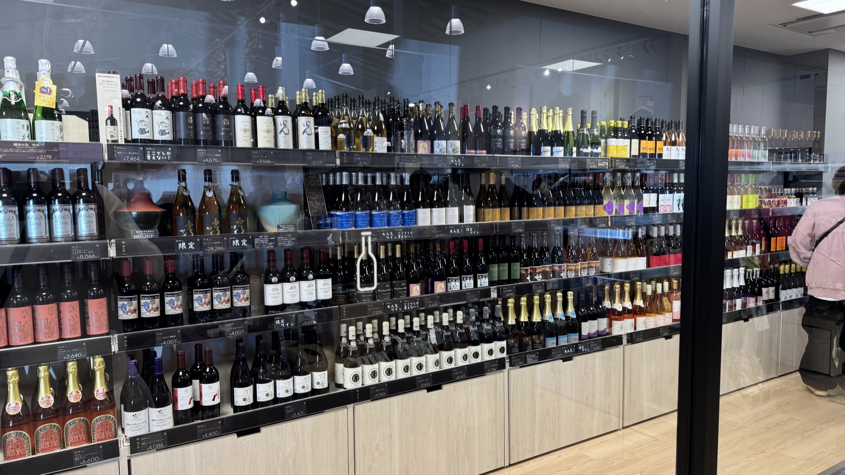 A large glass-fronted wall of Yamanashi wine and sake bottles with price tags at the Asama Market, Tabi-no-Eki Kawaguchiko Base.