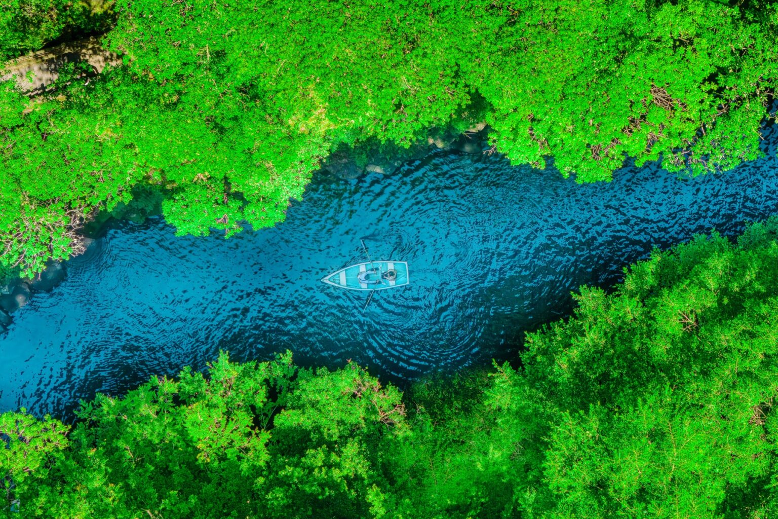An aerial top-down view of a single white rental rowboat on the teal river of Takachiho Gorge framed by bright green forest on both banks in Miyazaki, Kyushu.