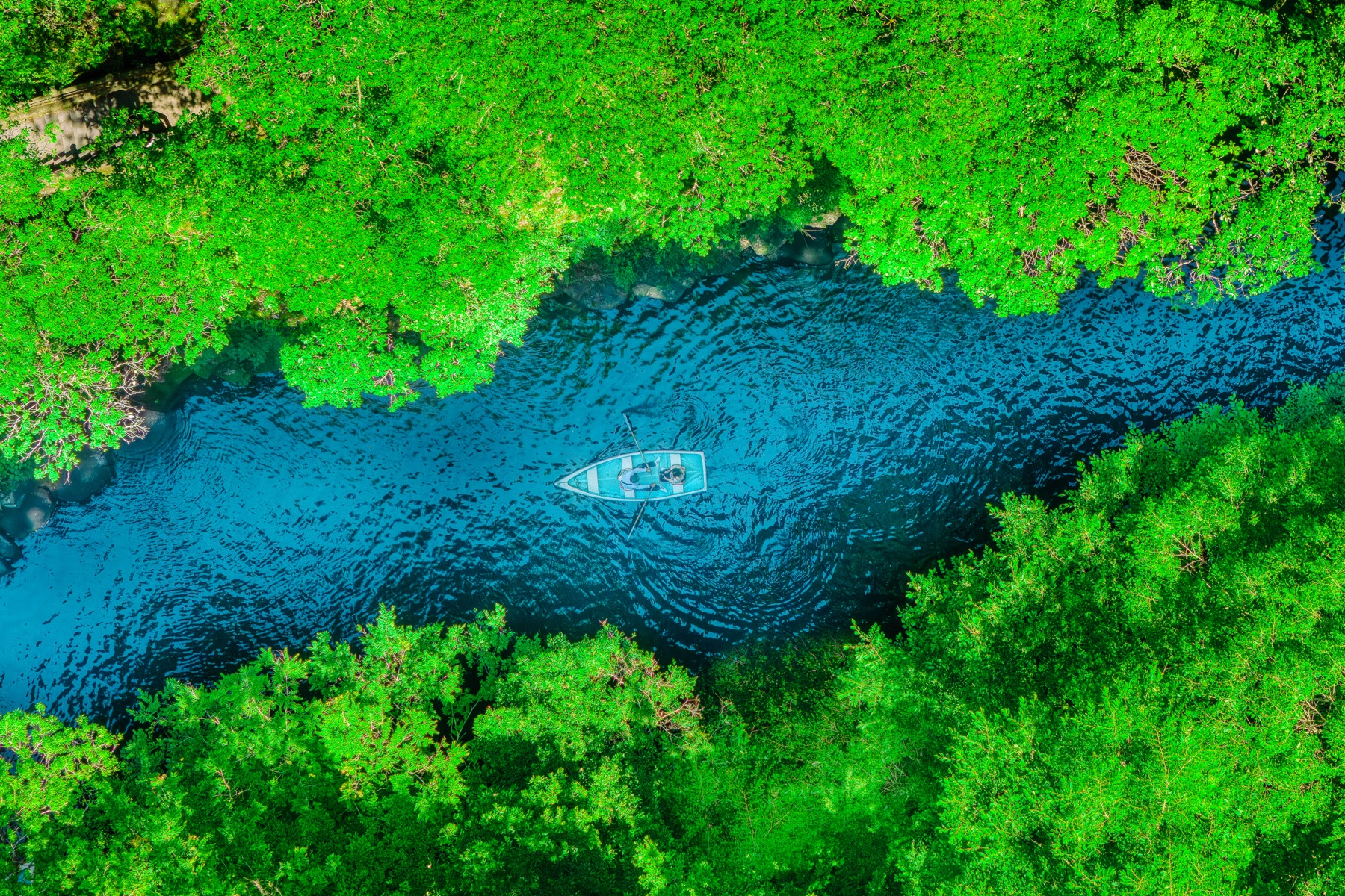 An aerial top-down view of a single white rental rowboat on the teal river of Takachiho Gorge framed by bright green forest on both banks in Miyazaki, Kyushu.