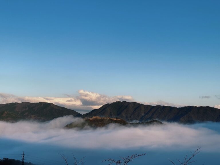 The stone ruins of Takeda Castle on a ridge above a thick sea of white clouds at dawn, with blue morning sky and dark mountains behind, seen from the Ritsuunkyo viewpoint in Hyogo.