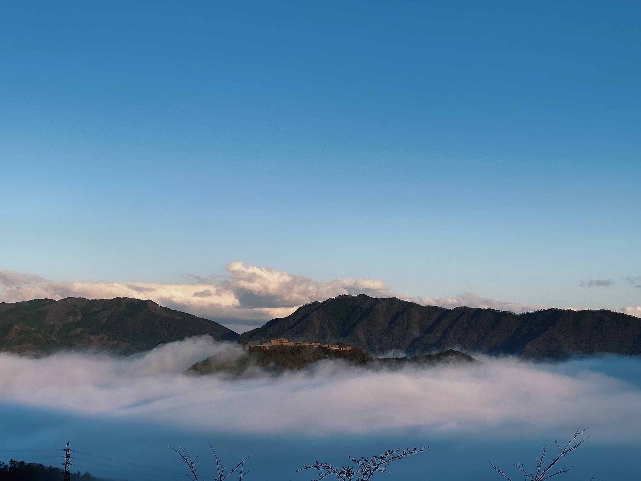 The stone ruins of Takeda Castle on a ridge above a thick sea of white clouds at dawn, with blue morning sky and dark mountains behind, seen from the Ritsuunkyo viewpoint in Hyogo.