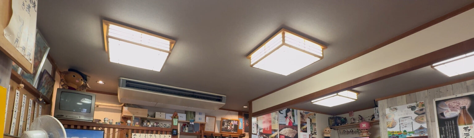 The interior of Takinoya soba shop in Mitaka: washi-paper ceiling lights, an old box TV on a shelf, hand-written menu strips on the wall and soba posters, in a wood-panelled room.