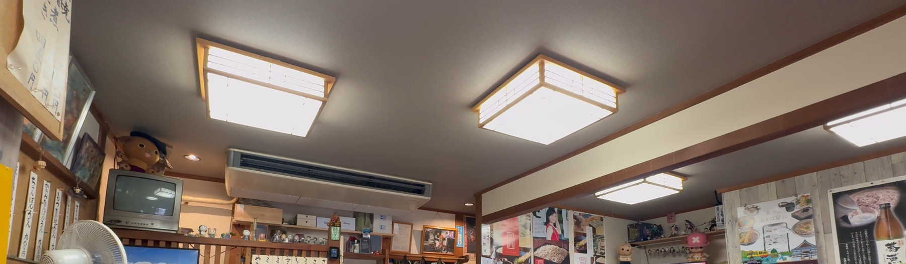 The interior of Takinoya soba shop in Mitaka: washi-paper ceiling lights, an old box TV on a shelf, hand-written menu strips on the wall and soba posters, in a wood-panelled room.