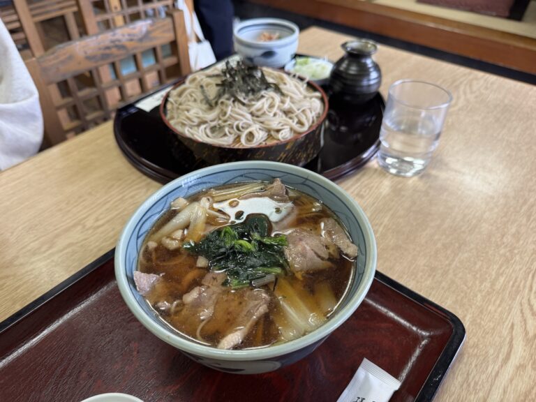 A duck soba set at Takinoya in Mitaka: a bowl of hot duck-and-leek dipping broth in front and a tray of chilled soba noodles behind, with condiments, on a wood table in the old soba shop.