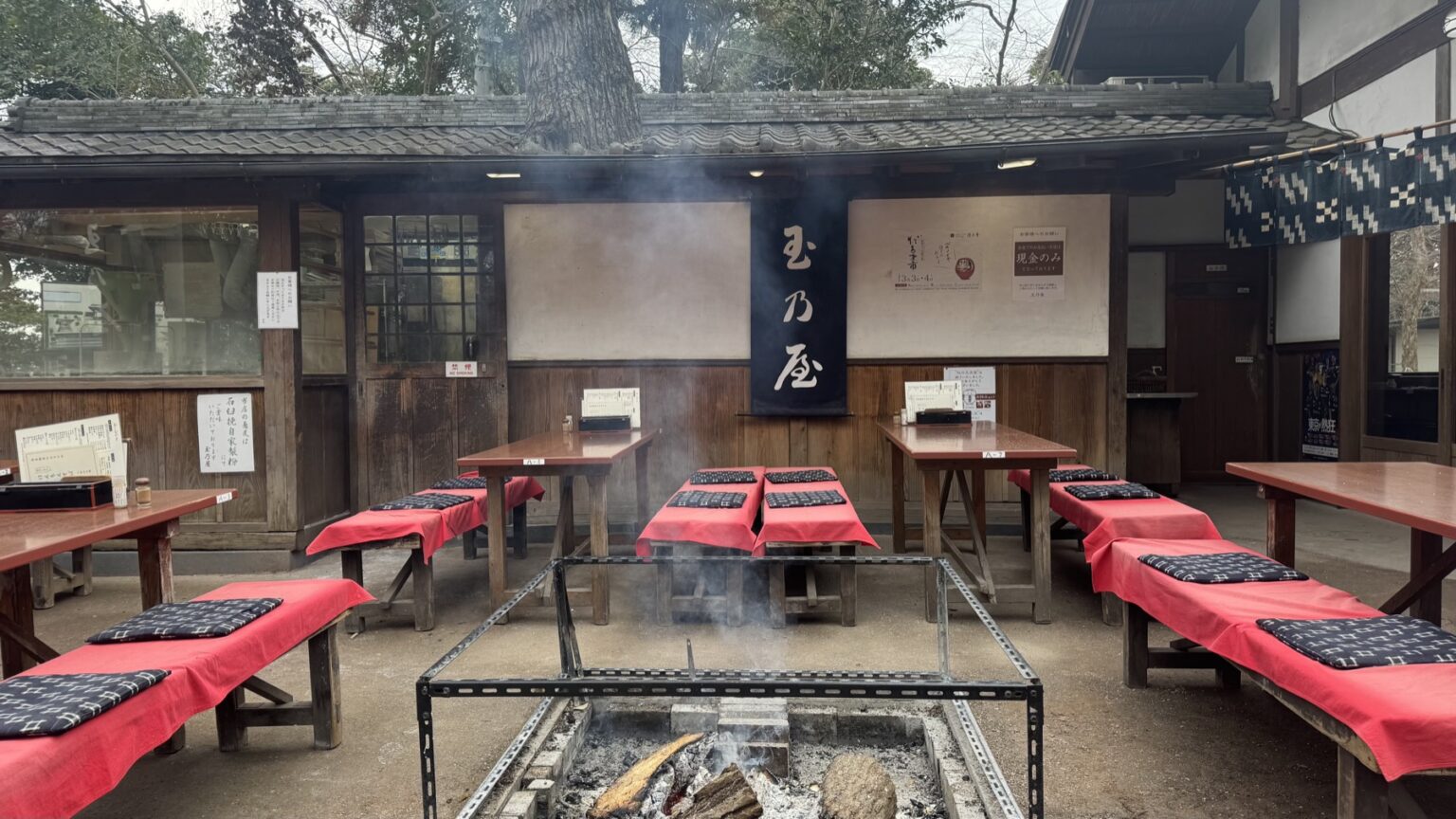 Outdoor bench seating with red cushions arranged around a sunken open charcoal hearth with smoke rising, in front of the wooden Tamanoya soba house in the temple town of Jindaiji, Tokyo