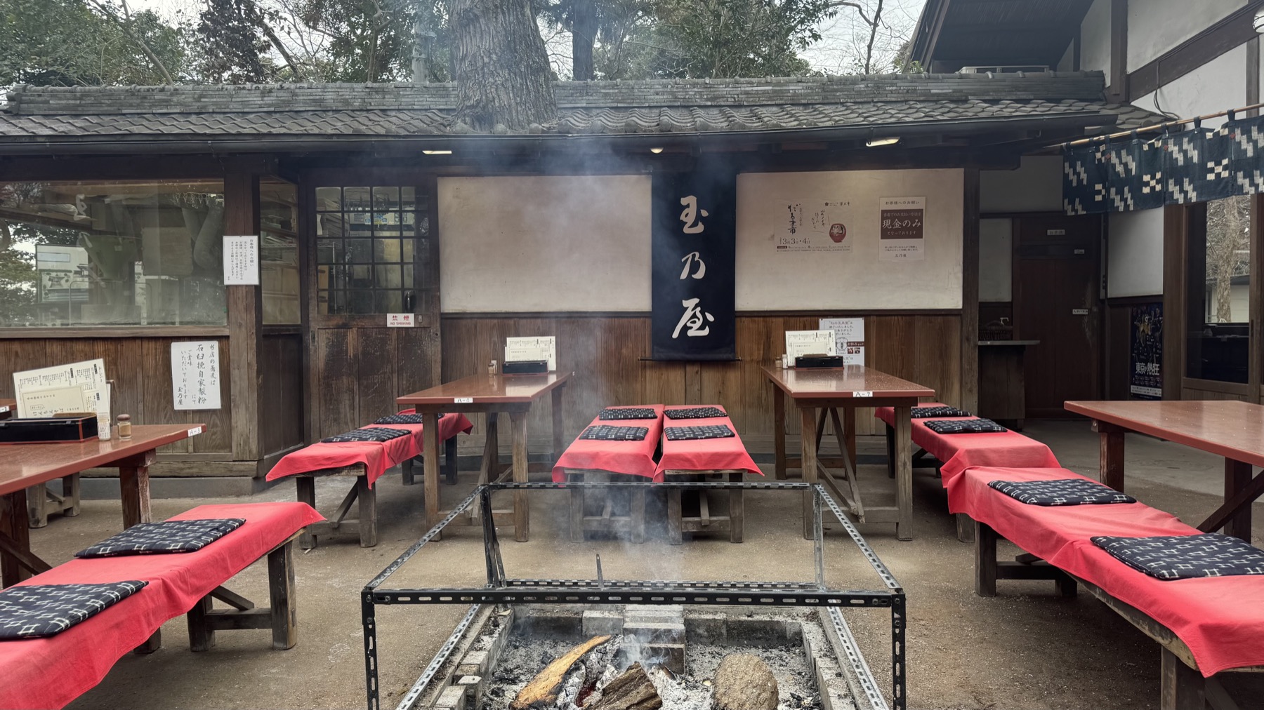 Outdoor bench seating with red cushions arranged around a sunken open charcoal hearth with smoke rising, in front of the wooden Tamanoya soba house in the temple town of Jindaiji, Tokyo