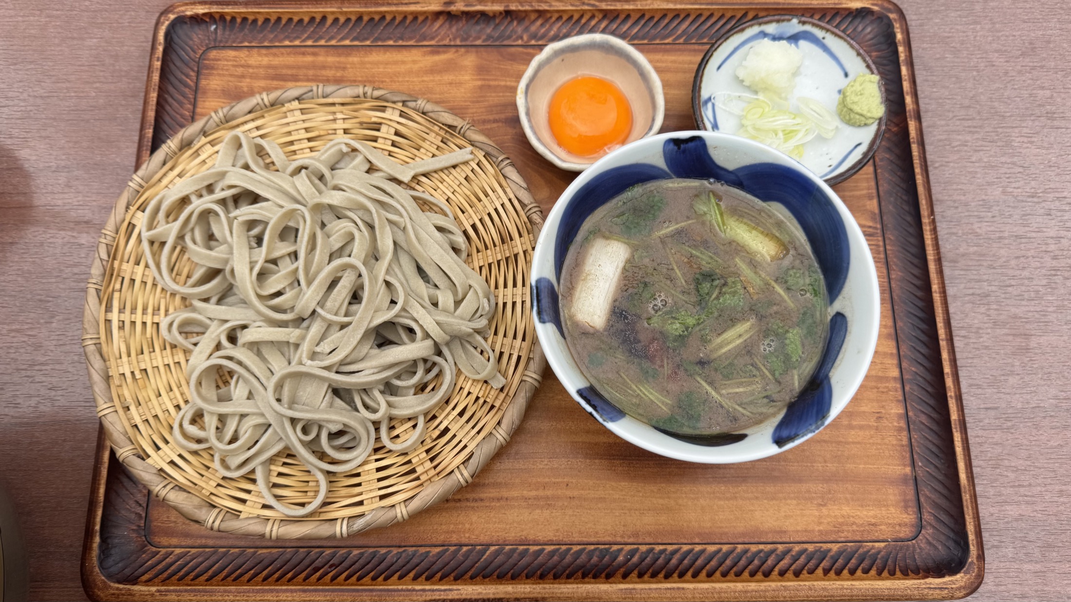 A bamboo basket of dark, hand-cut juwari buckwheat soba beside a bowl of hot duck-and-leek dipping broth with wasabi and chopped leek on a wooden tray at a Tokyo soba house