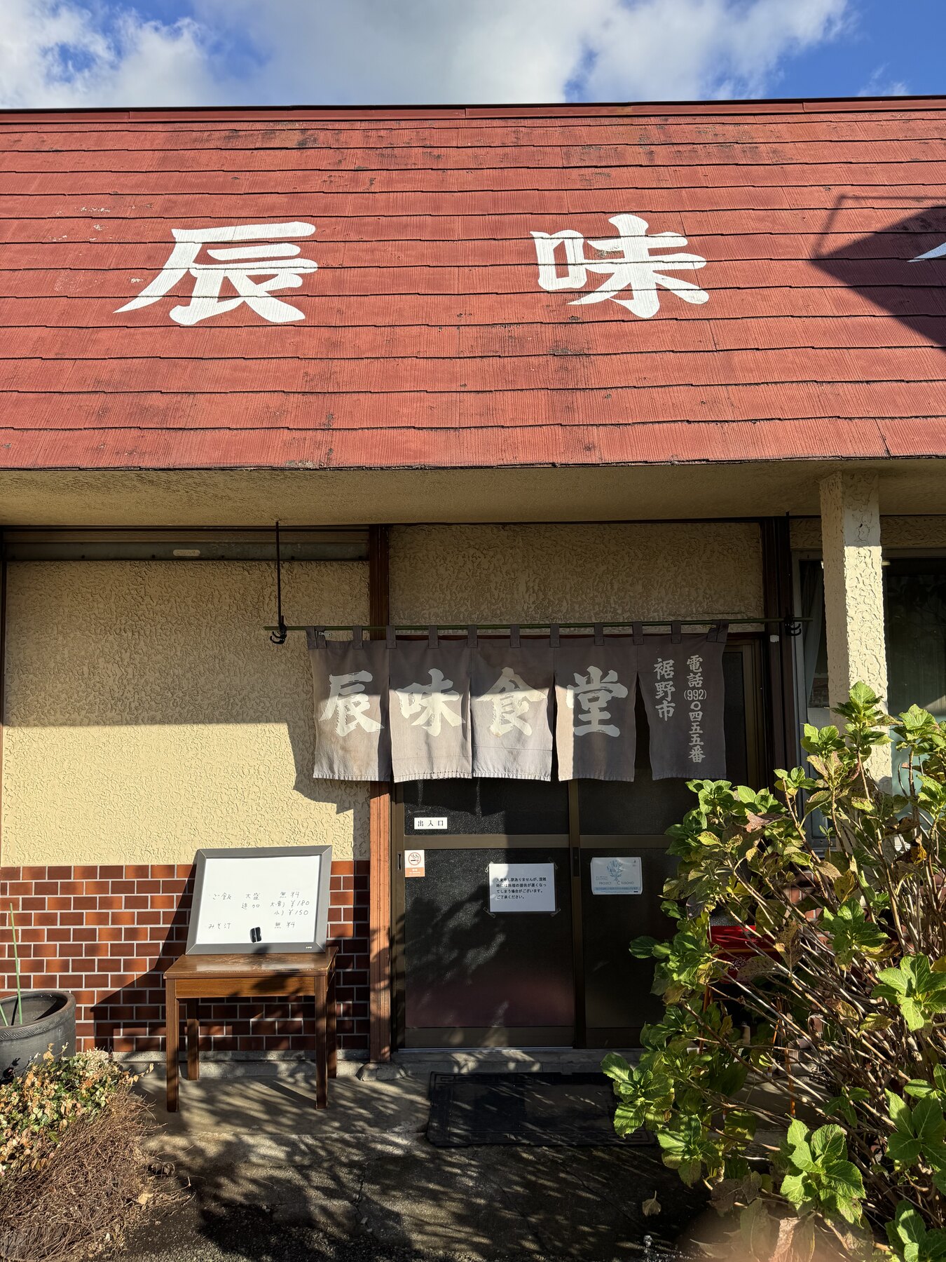 The entrance of Tatsumi Shokudo in Susono with its red wall, white noren curtain and a hand-written menu board beside the door, framed by green leaves.