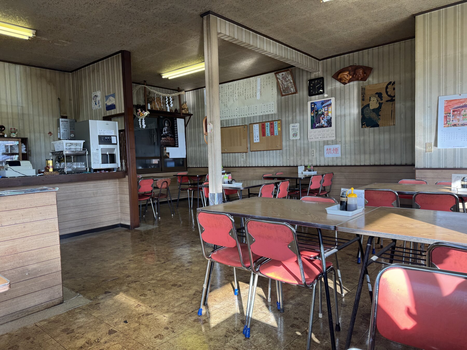 The retro interior of Tatsumi Shokudo in Susono: wood-panelled walls, red vinyl chairs around formica tables, fluorescent lights and old trophies on a shelf by the kitchen counter.