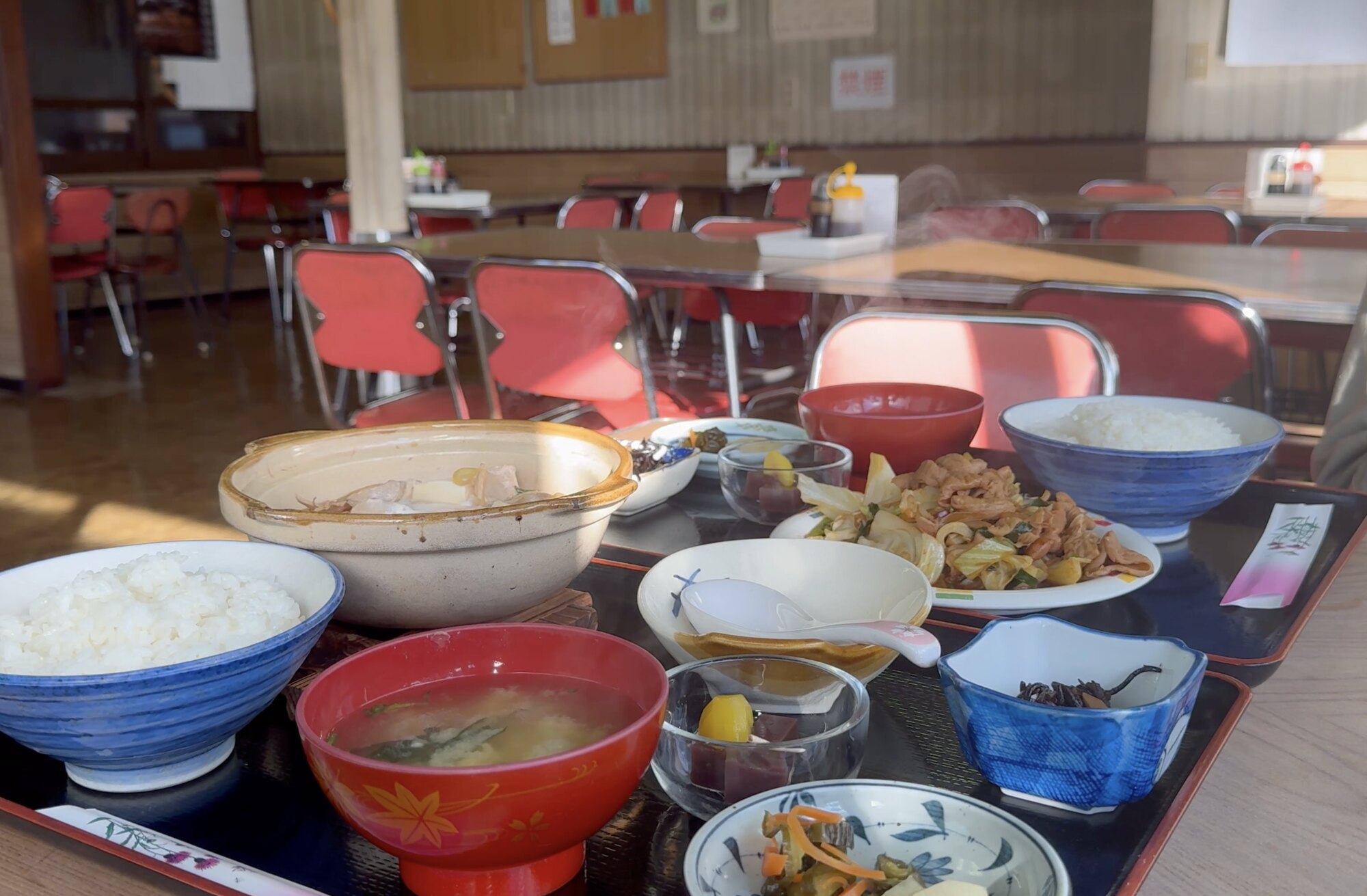 Two teishoku set meals on a table at Tatsumi Shokudo in Susono: two large bowls of white rice, a clay pot of stew, red bowls of miso soup and small dishes of stewed meat, vegetables, pickles and egg, with the retro dining room behind.