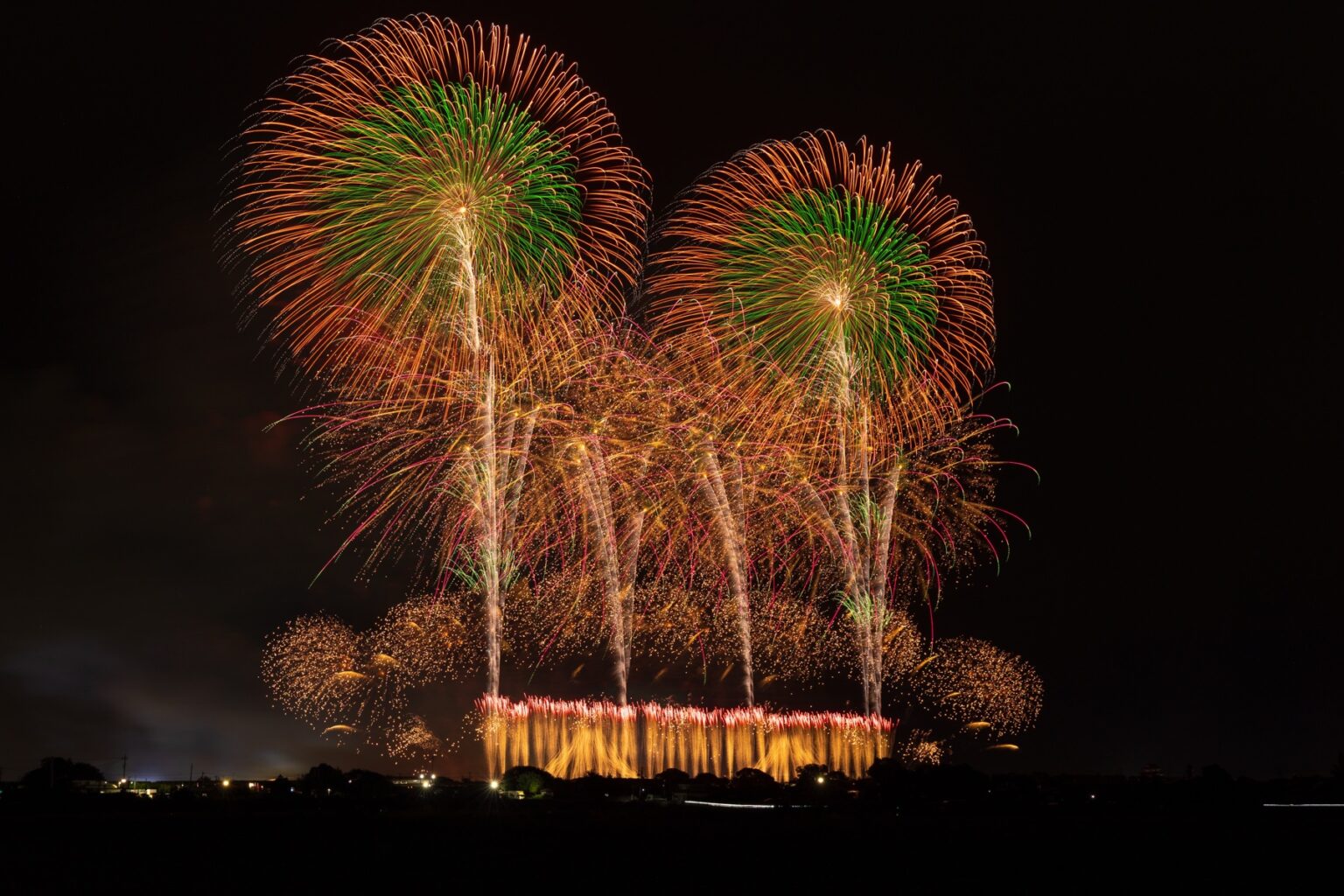 Two enormous chrysanthemum fireworks shells bloom side by side, shifting from green centres to orange tips, above a bright golden star-mine along the Tone River bank.