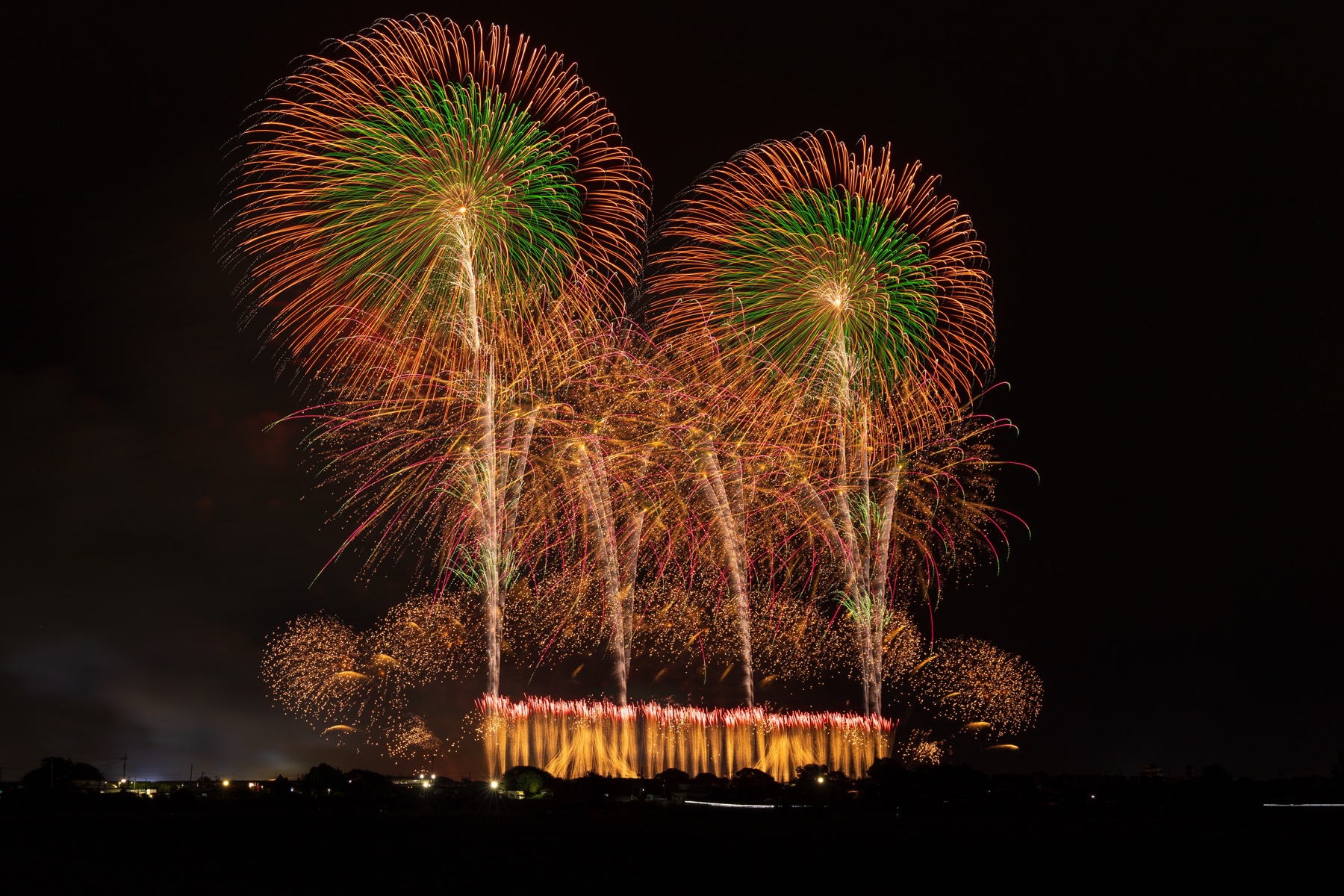Two enormous chrysanthemum fireworks shells bloom side by side, shifting from green centres to orange tips, above a bright golden star-mine along the Tone River bank.