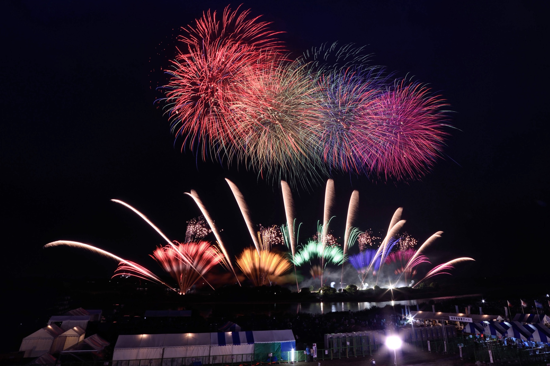 Large colourful fireworks burst above rows of festival tents and spectators lining the Tone River bank, with reflections shimmering on the water below.