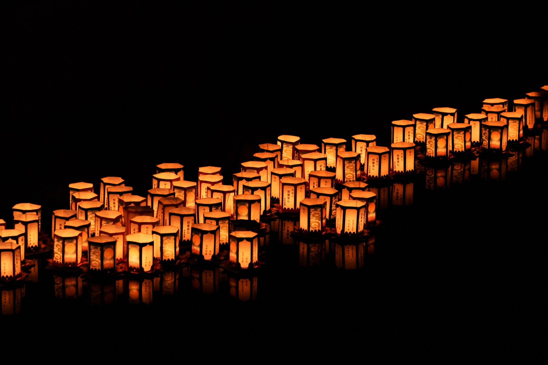 Rows of glowing paper toro-nagashi lanterns floating on dark still water at night during Obon in Japan, their warm orange light reflected on the surface as they drift to send off ancestral spirits