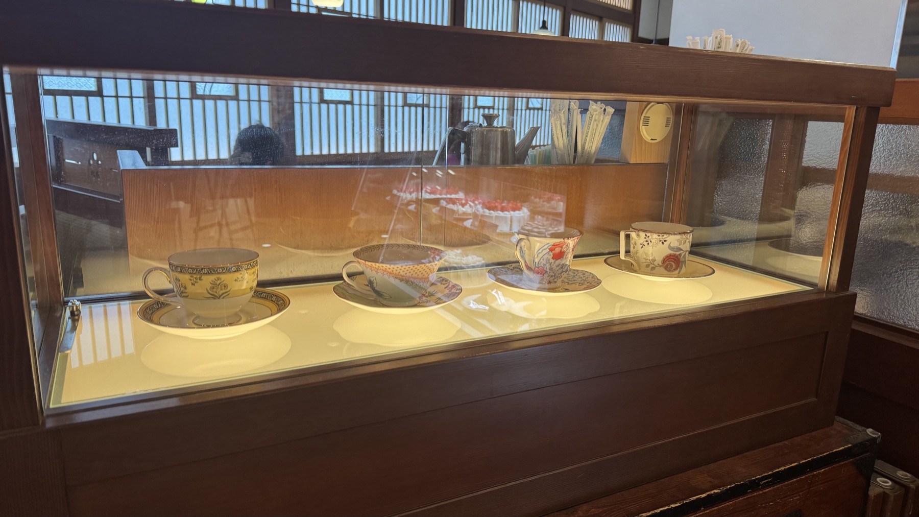 A wooden-framed glass display case at Tsubakiya Cafe Kichijoji showing a row of antique-style coffee cups and saucers in different floral patterns, the kind used to serve coffee at the table