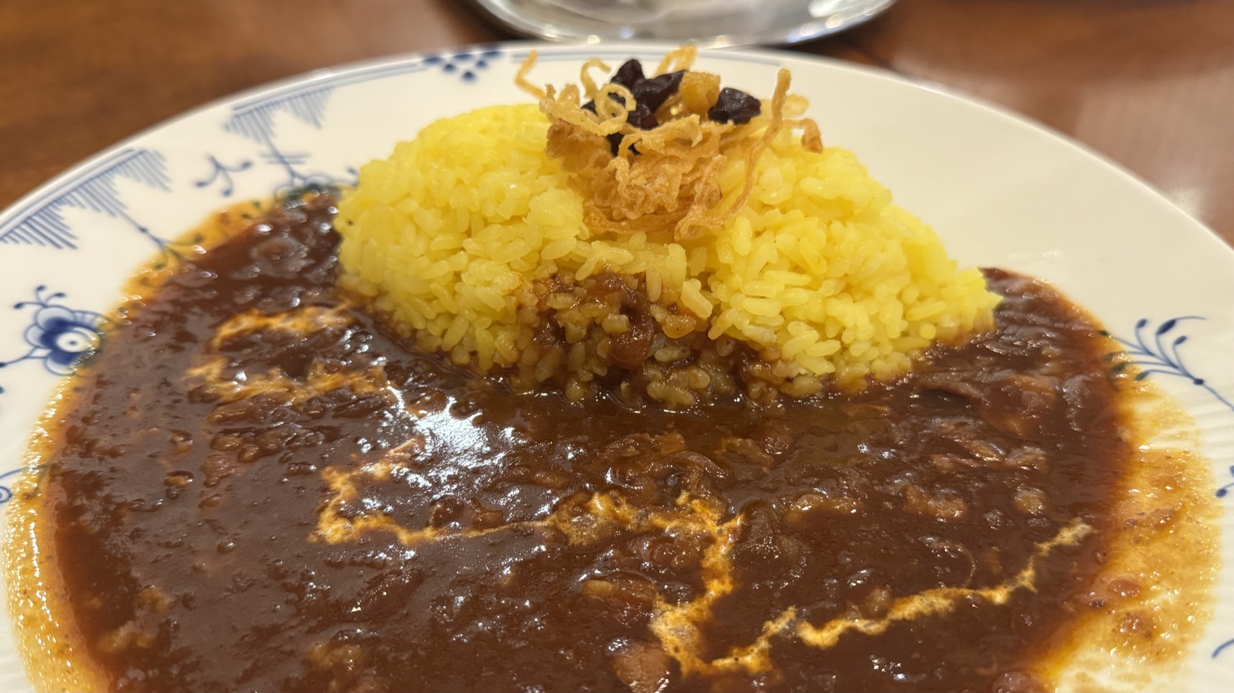 A close-up of Tsubakiya Cafe's beef curry, glossy dark long-simmered sauce around a mound of bright yellow turmeric rice topped with crisp fried onions, served on a blue-and-white patterned plate