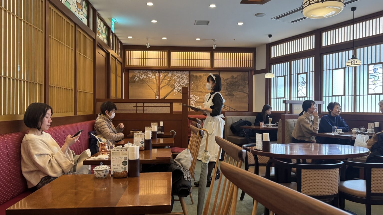 The Taisho-roman interior of Tsubakiya Cafe Kichijoji with wooden kumiko lattice, stained glass, a painted cherry-blossom screen and pink banquette seats, and a waitress in a classic black dress and white apron serving a table
