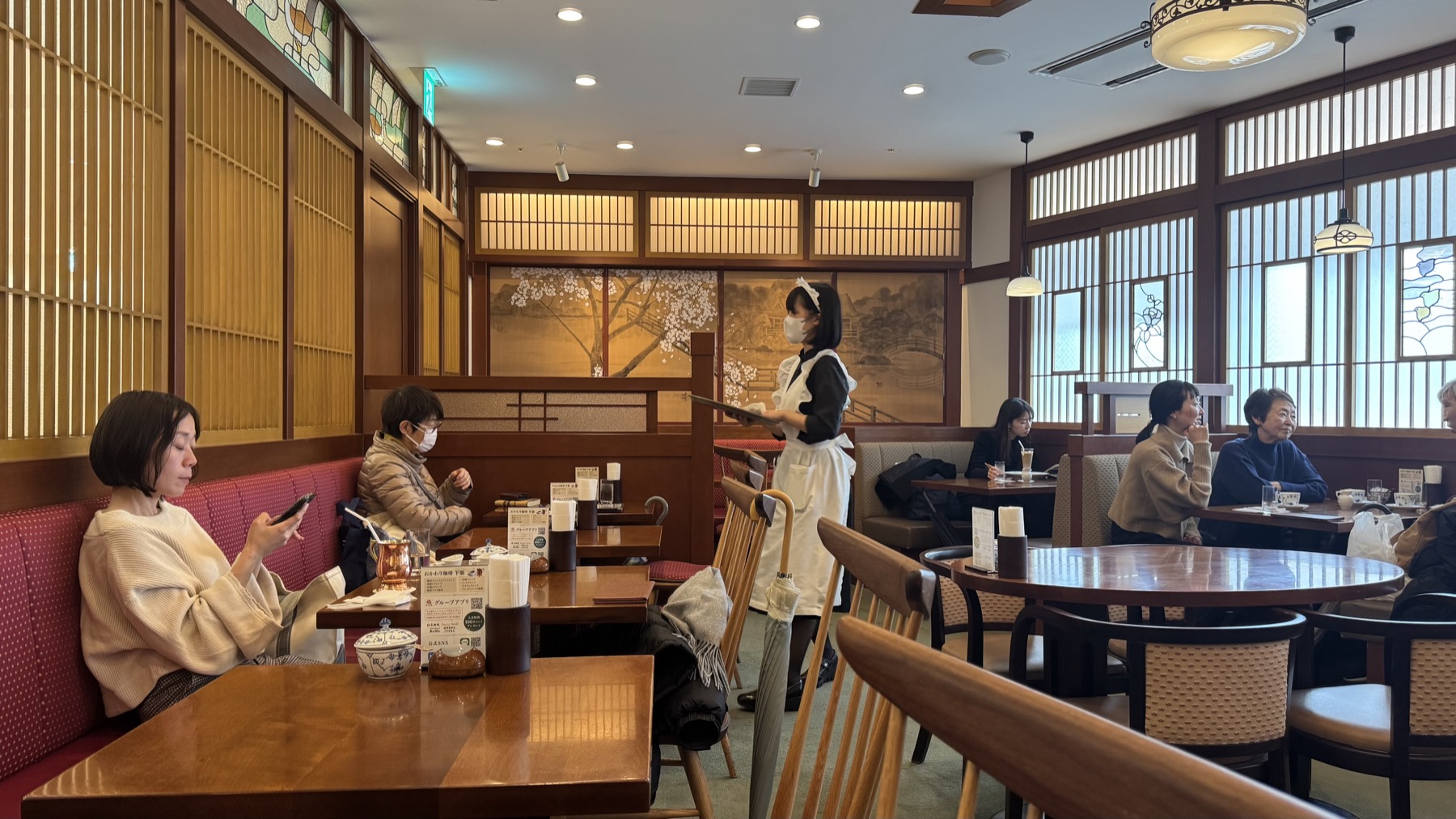 The Taisho-roman interior of Tsubakiya Cafe Kichijoji with wooden kumiko lattice, stained glass, a painted cherry-blossom screen and pink banquette seats, and a waitress in a classic black dress and white apron serving a table
