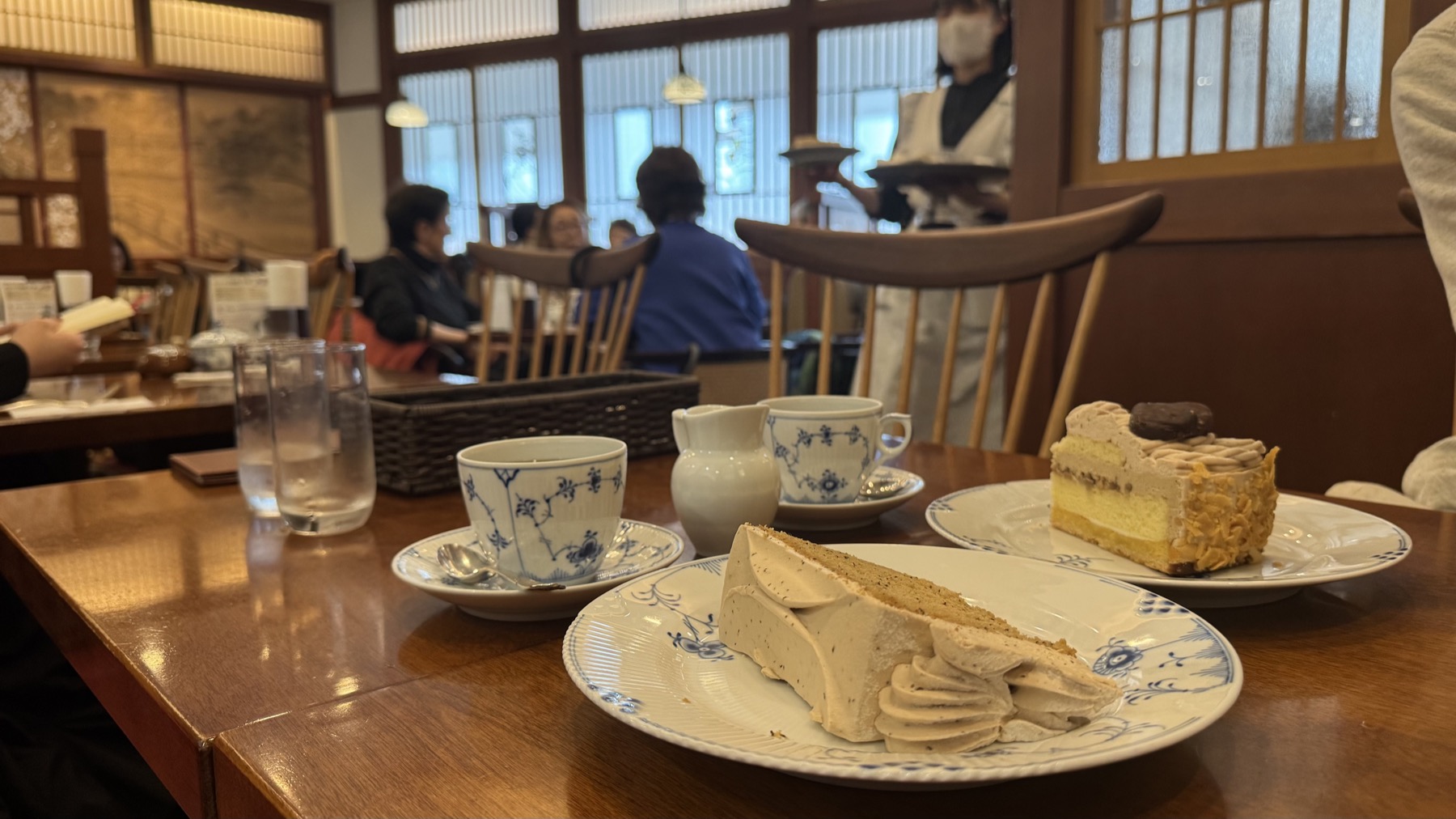 Two slices of chiffon cake and two cups of coffee in Royal Copenhagen blue-fluted cups at Tsubakiya Cafe Kichijoji, with water glasses and a milk jug, the retro cafe room and a uniformed waitress behind