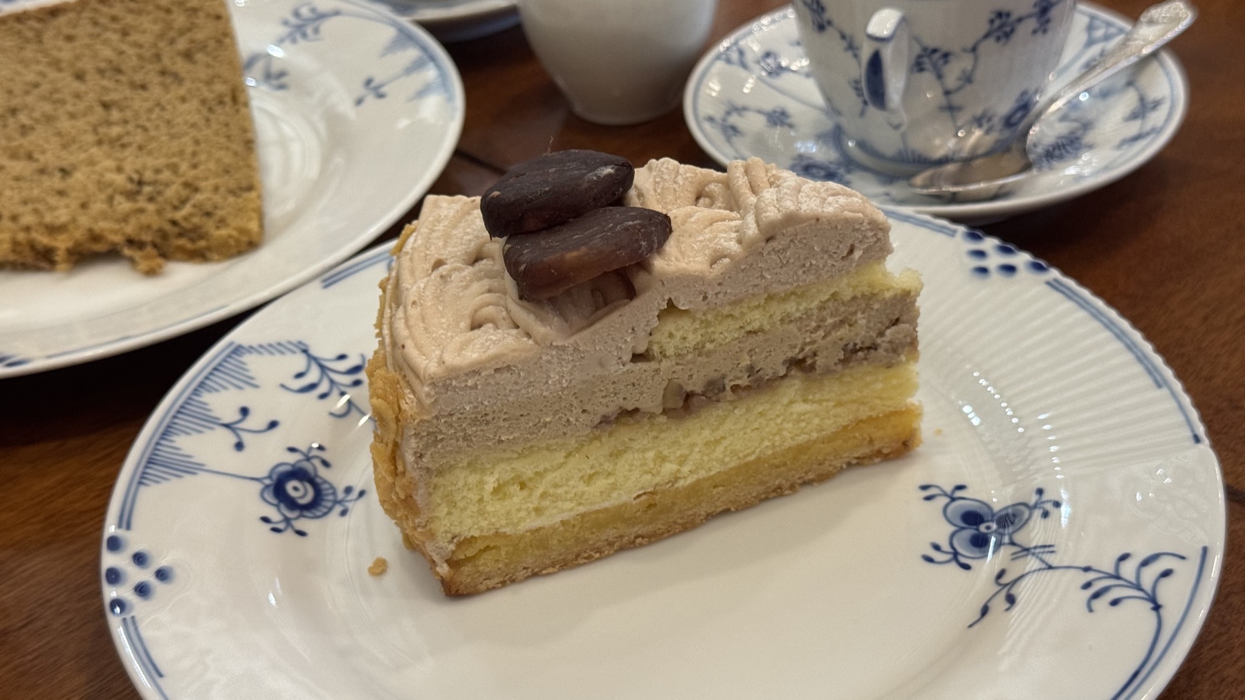 A close-up slice of layered chiffon cake at Tsubakiya Cafe, light sponge with coffee-brown cream and chocolate beans on top, on a blue-fluted plate, with a plain chiffon slice and a coffee cup behind