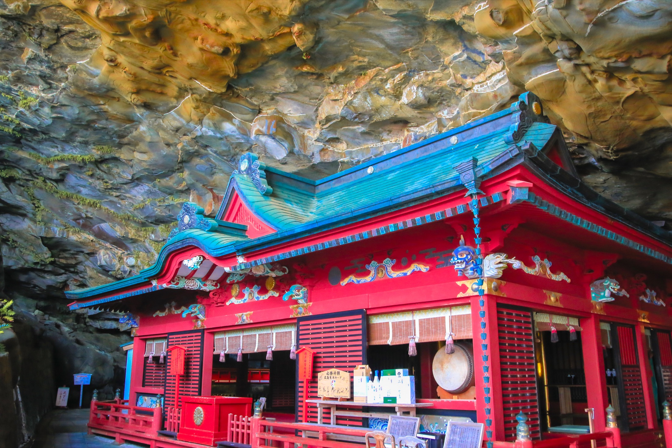 The vermilion and turquoise main hall of Udo Jingu shrine standing inside a large coastal rock cave, its ornate roof beneath the cave ceiling on the Nichinan coast of Miyazaki.