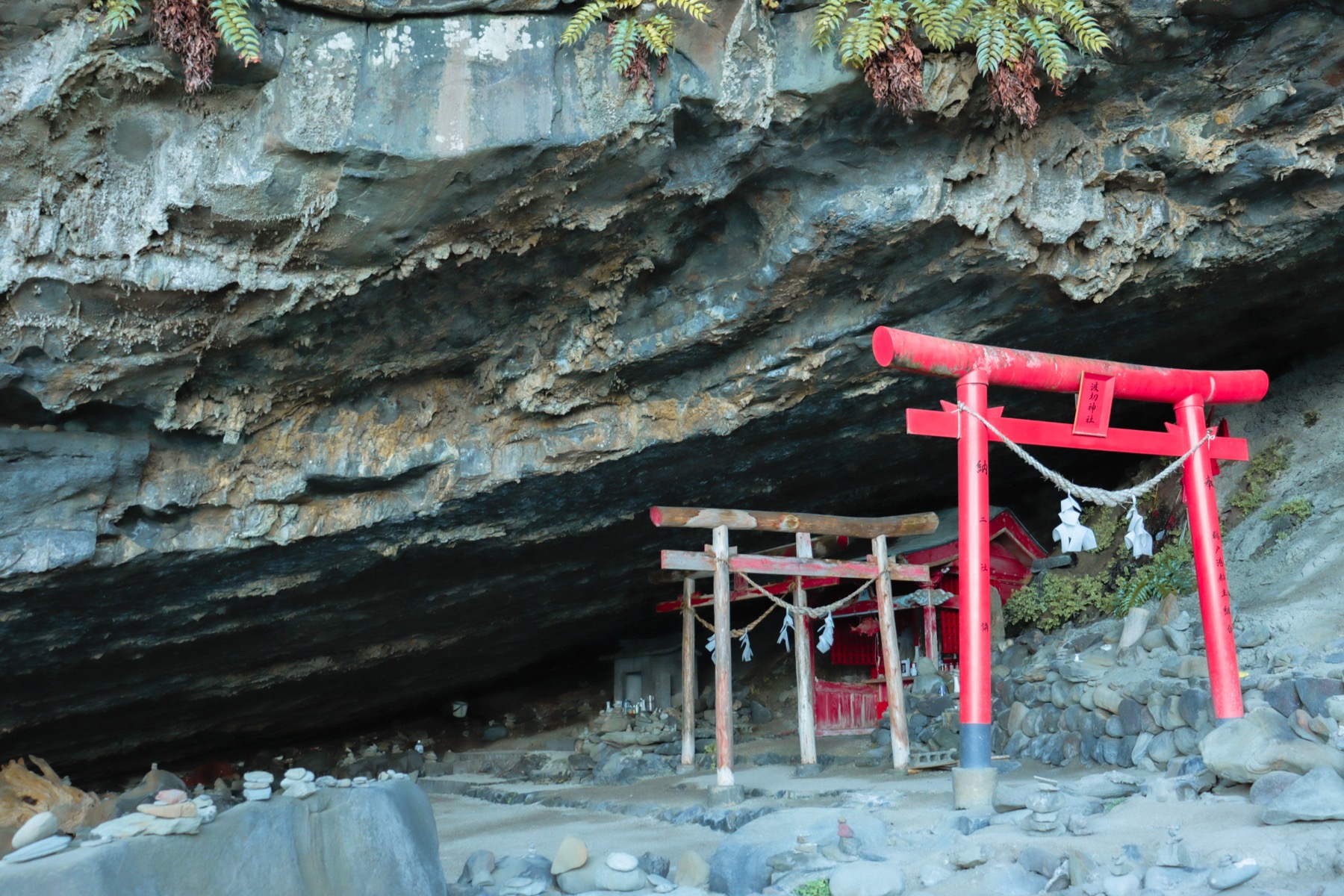 A small red torii and tiny shrine with stacked prayer stones tucked under a sea-cave overhang at Udo Jingu on the Nichinan coast, Miyazaki.
