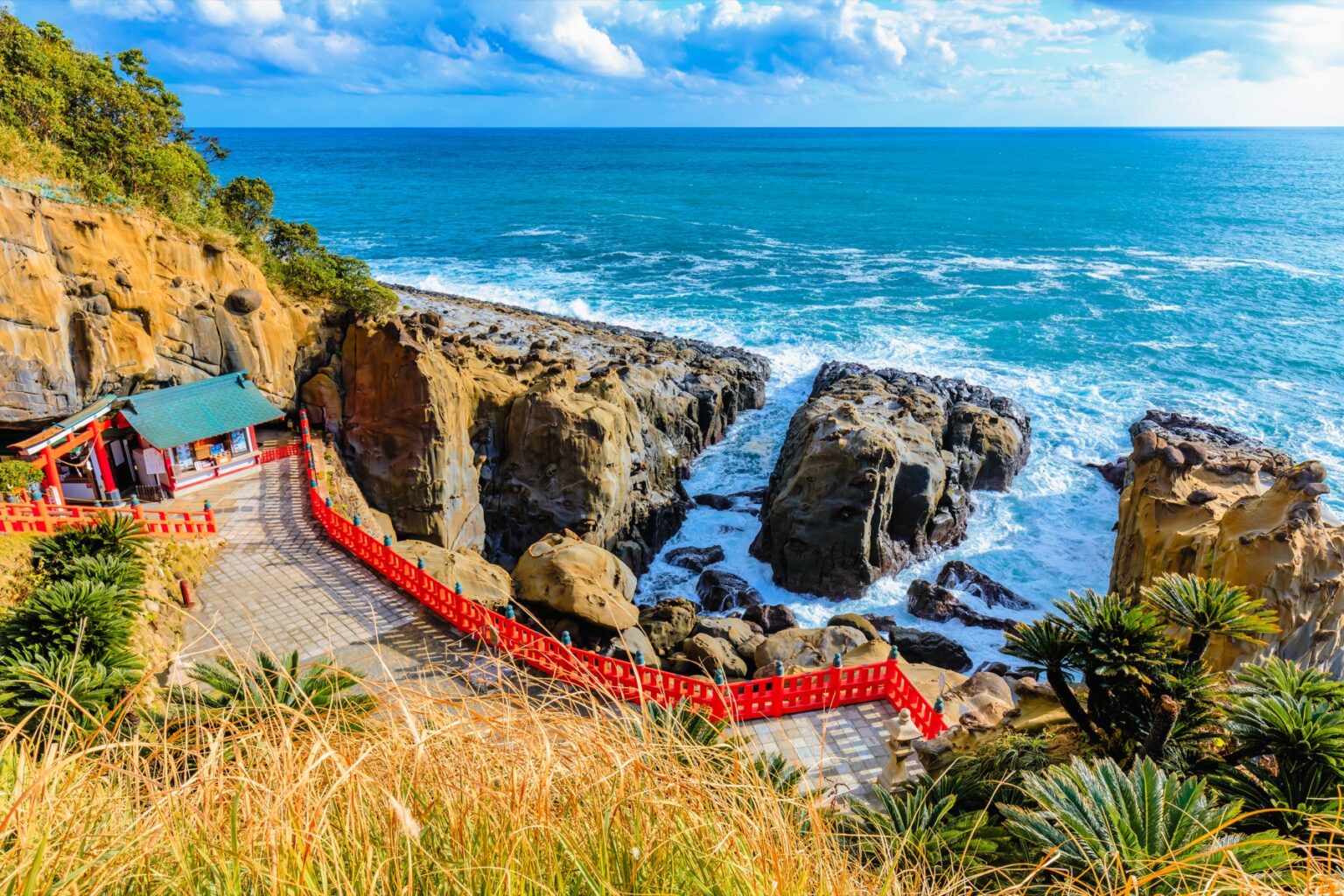 The vermilion buildings and red railings of Udo Jingu tucked at the foot of a cliff above the rocky Hyuga-nada coast, the blue sea and surf beyond, in Nichinan, Miyazaki.