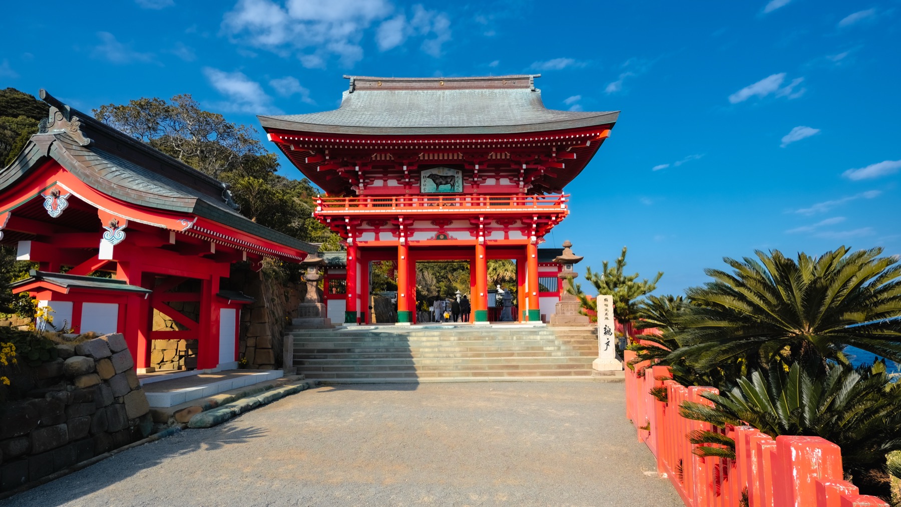 The grand vermilion tower gate of Udo Jingu shrine at the top of stone steps flanked by palms under a blue sky on the Nichinan coast of Miyazaki.